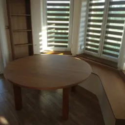 Okrągły, drewniany stół z trzema nogami w jasnym pomieszczeniu z oknami z żaluzjami poziomymi i wbudowaną szafką z półkami.