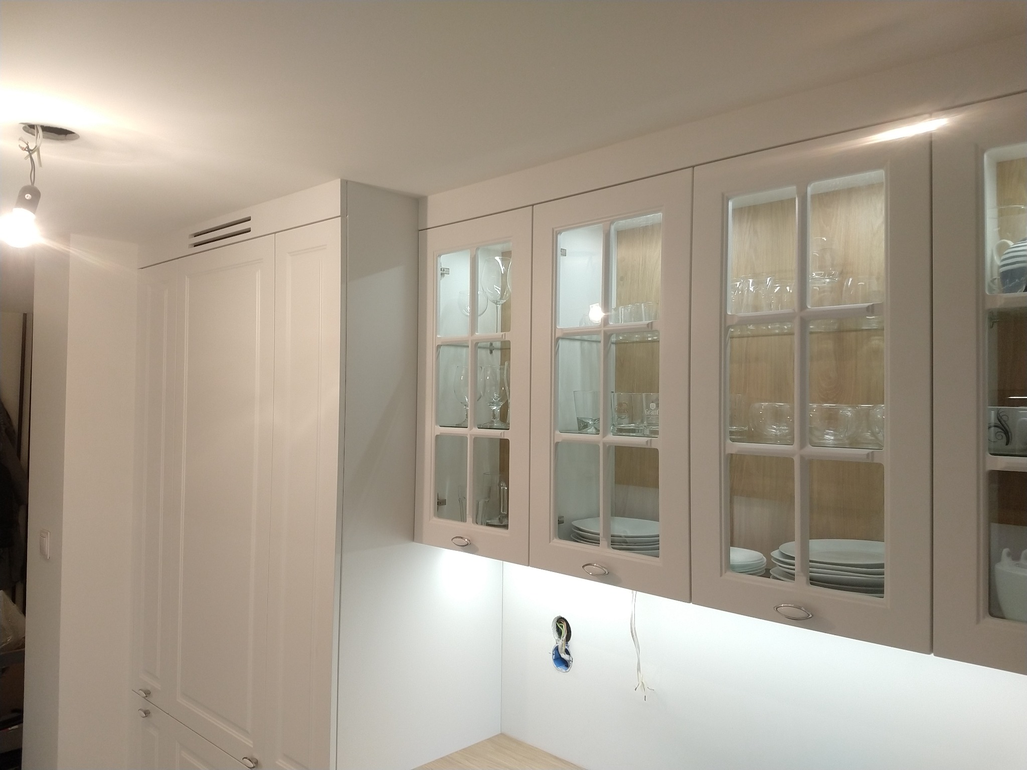 Białe, górne szafki kuchenne z przeszklonymi frontami, oświetleniem LED podszafkowym i widoczną instalacją elektryczną w ścianie.
