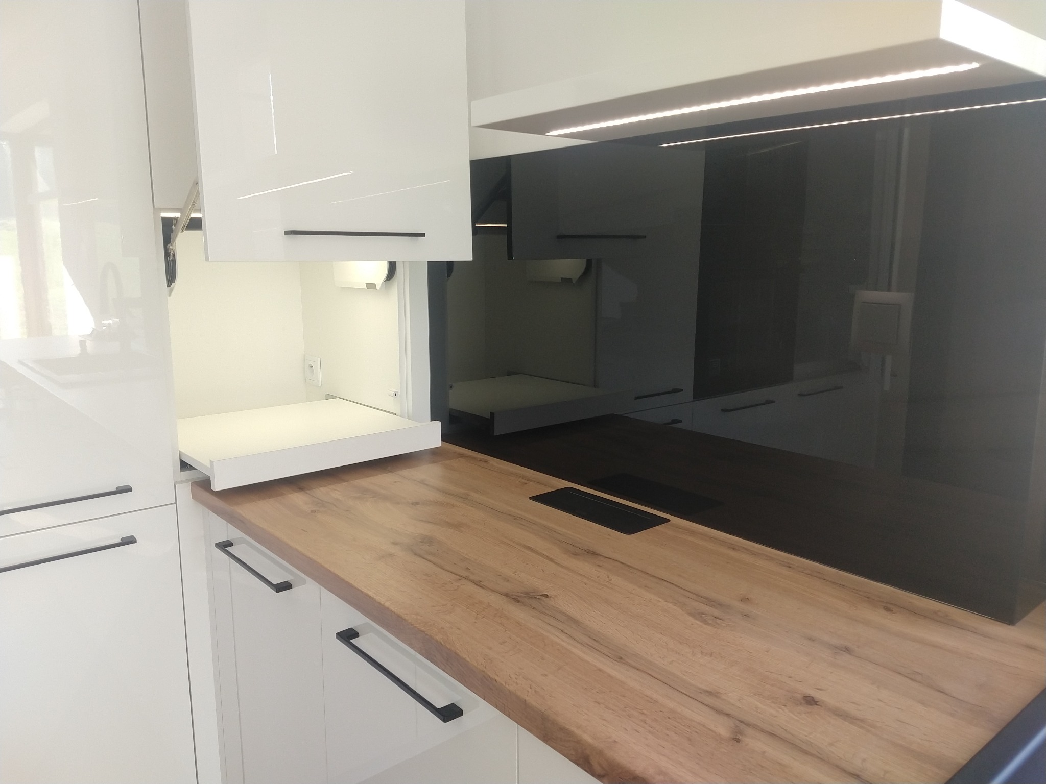 Nowoczesna kuchnia z białymi, lakierowanymi szafkami i drewnianym blatem z wbudowanym gniazdkiem, oświetlenie LED pod szafkami wiszącymi.