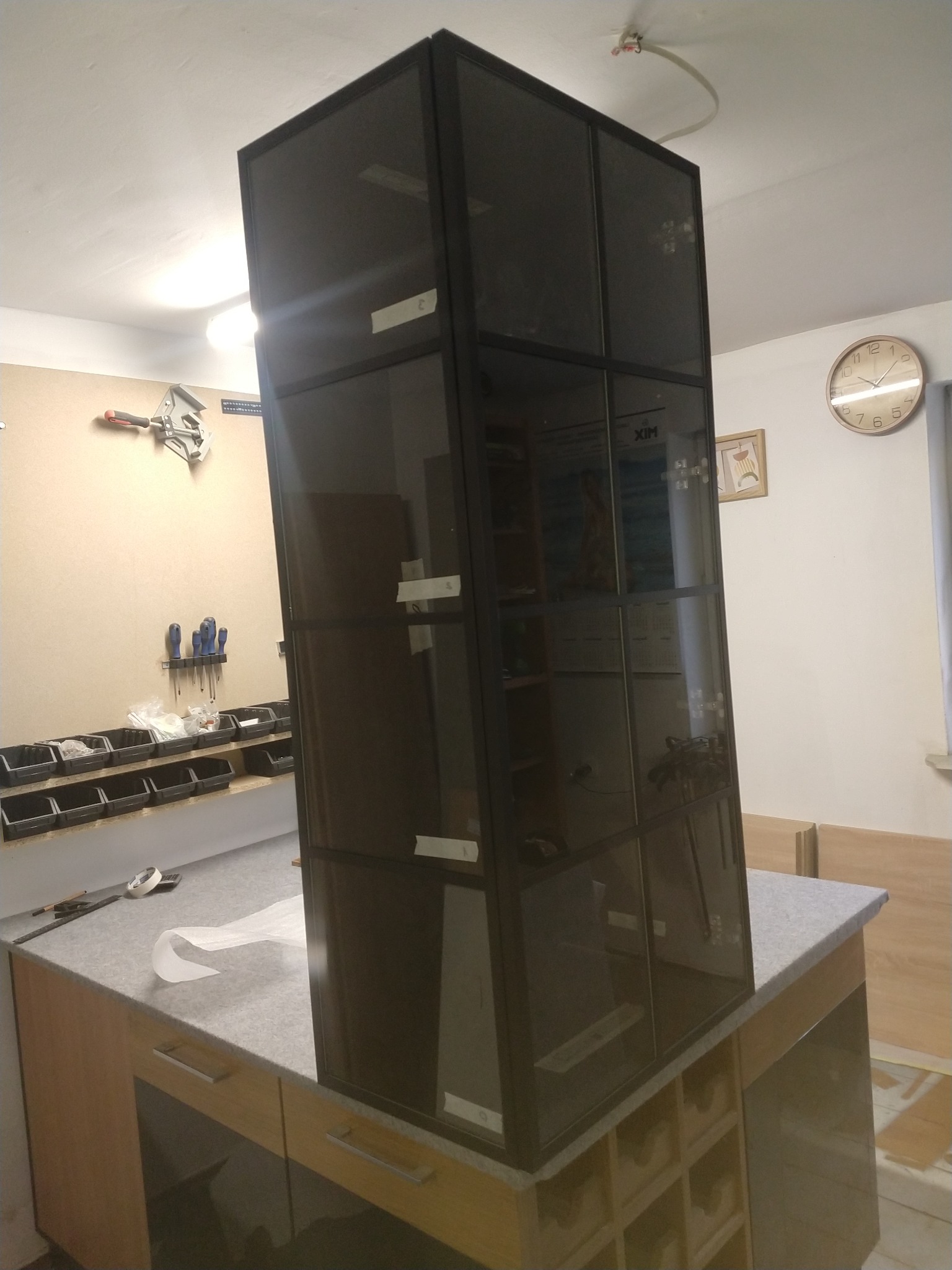 Wysoka, przeszklona szafa z czarną ramą, tymczasowo zabezpieczona białymi taśmami, stoi na blacie roboczym w warsztacie. Widoczne narzędzia na ścianie i zegar.