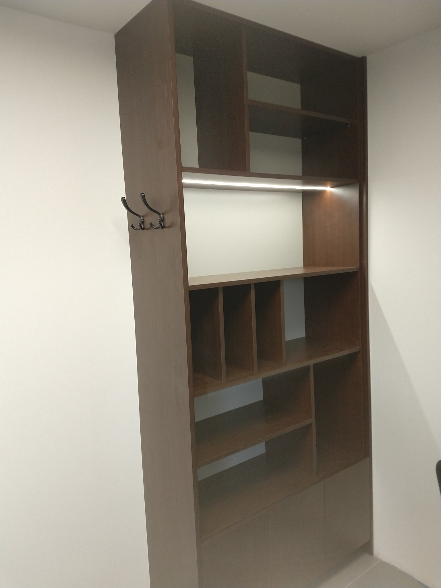 Wysoka, brązowa szafa wnękowa z oświetleniem LED i wieszakami na ubrania zamontowanymi z boku, widoczna na tle białej ściany.