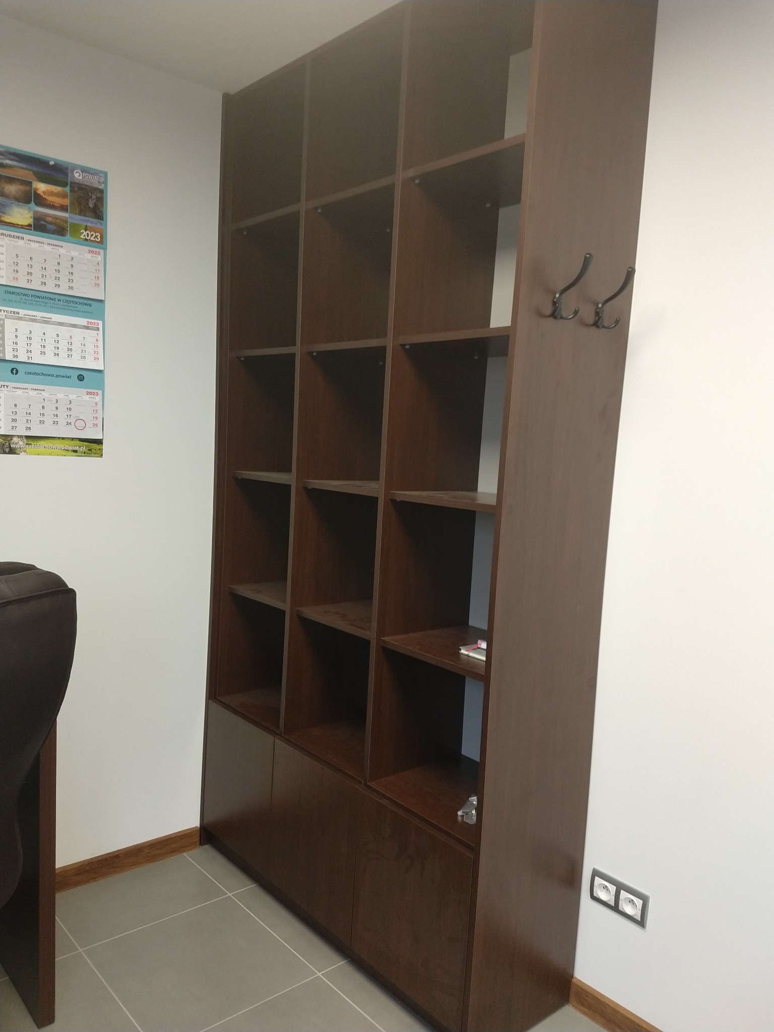Wysoka, brązowa szafa z otwartymi półkami i dolnymi szafkami, uzupełniona o dwa haczyki na ubrania po prawej stronie, widoczna na tle białej ściany z kalendarzem i gniazdkiem elektrycznym.