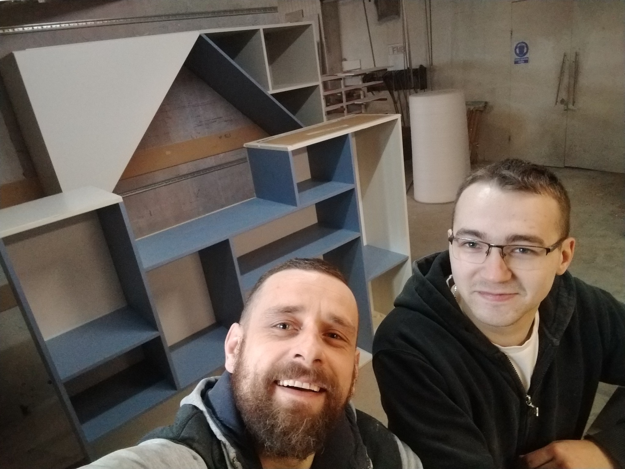 Zdjęcie selfie dwóch uśmiechniętych mężczyzn na tle elementów mebli w trakcie montażu w warsztacie, widoczne półki i ścianki w kolorach białym i szarym.