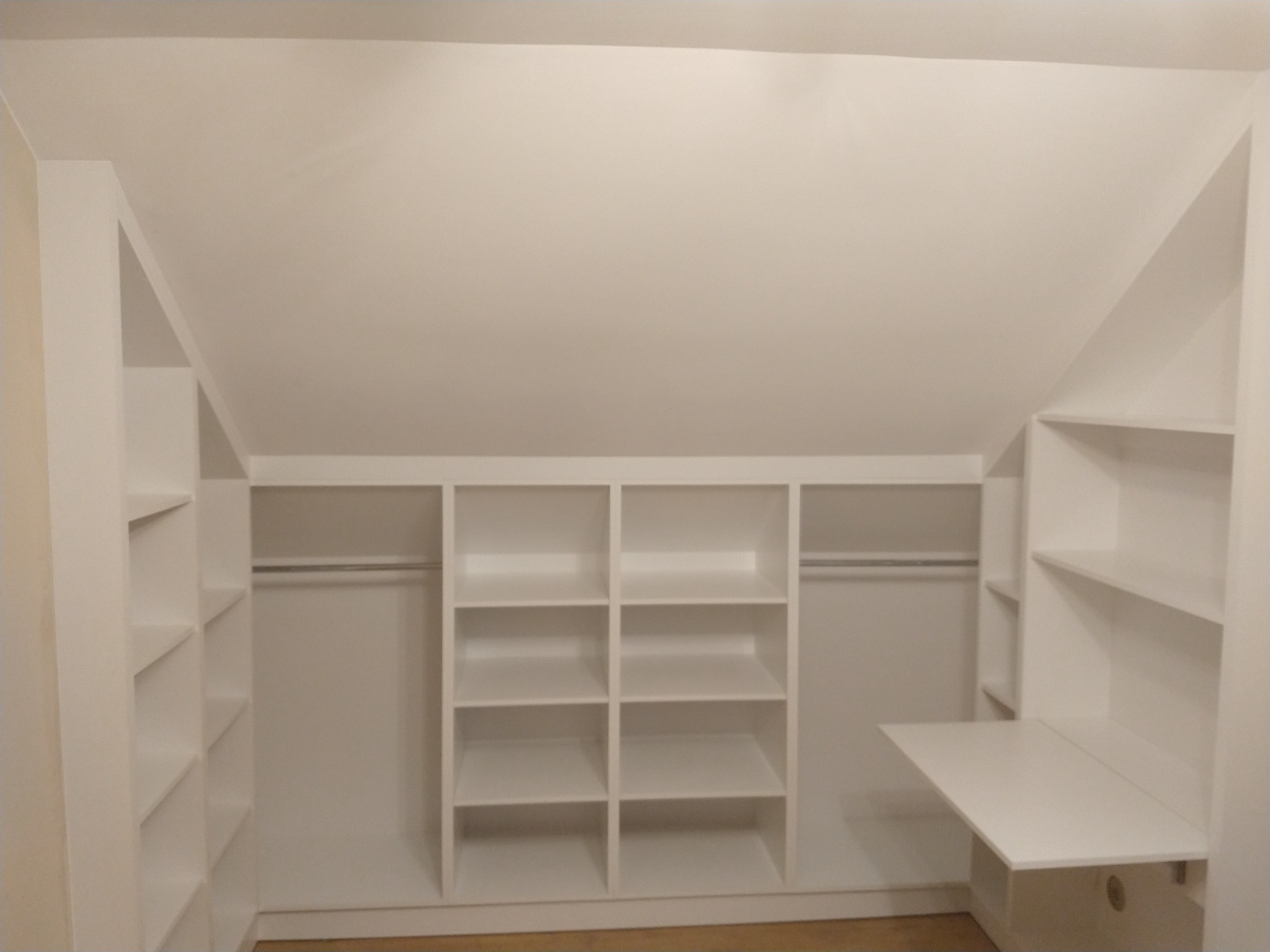 Biała szafa wnękowa z półkami i drążkami na ubrania, zabudowana na poddaszu ze skosem, z dodatkowym rozkładanym blatem biurka po prawej stronie.