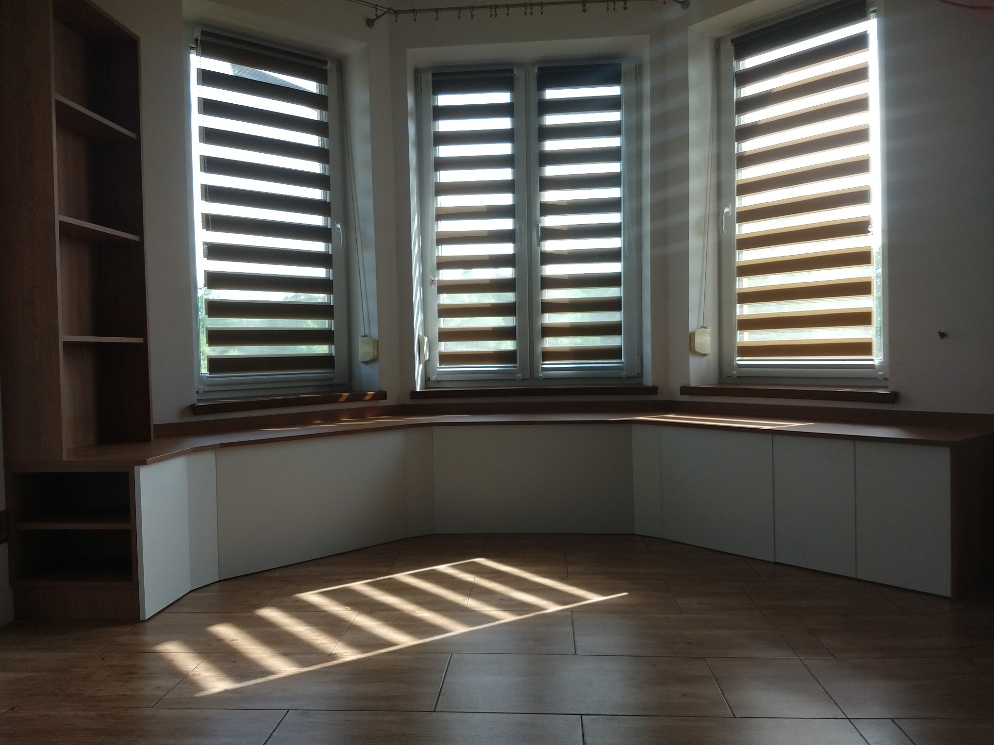 Wnęka okienna z zabudową meblową: szafka z blatem z drewna, obok regał, na podłodze płytki imitujące drewno, promienie słońca wpadające przez okno z roletami dzień-noc.