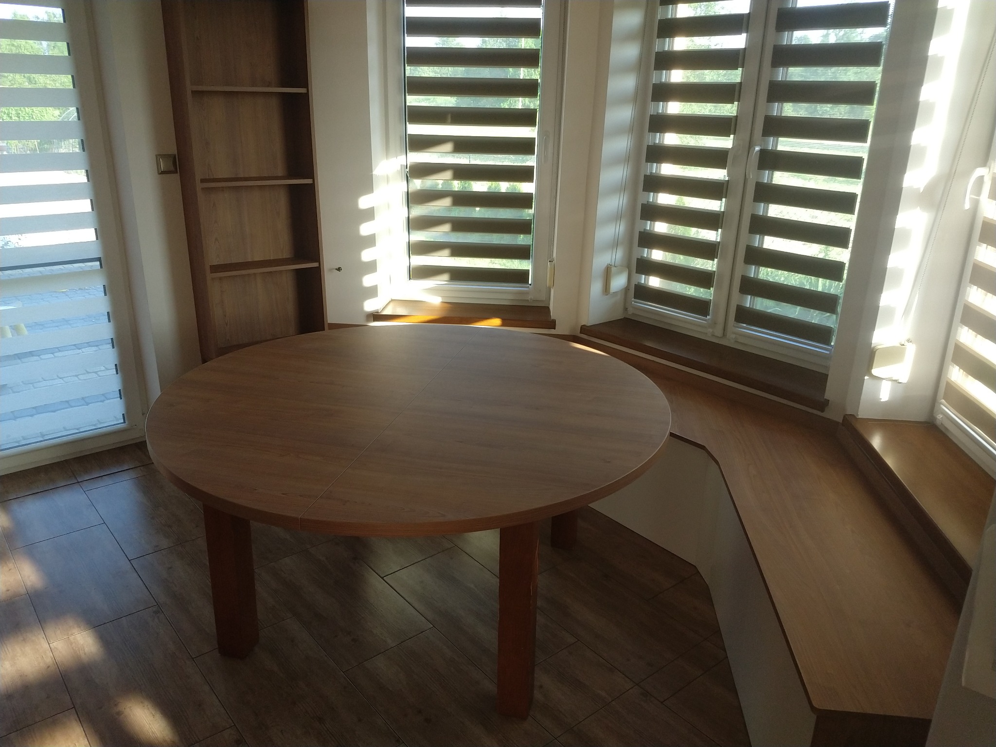 Okrągły, drewniany stół z trzema nogami w jasnym pomieszczeniu z oknami z żaluzjami poziomymi i wbudowaną szafką z półkami.