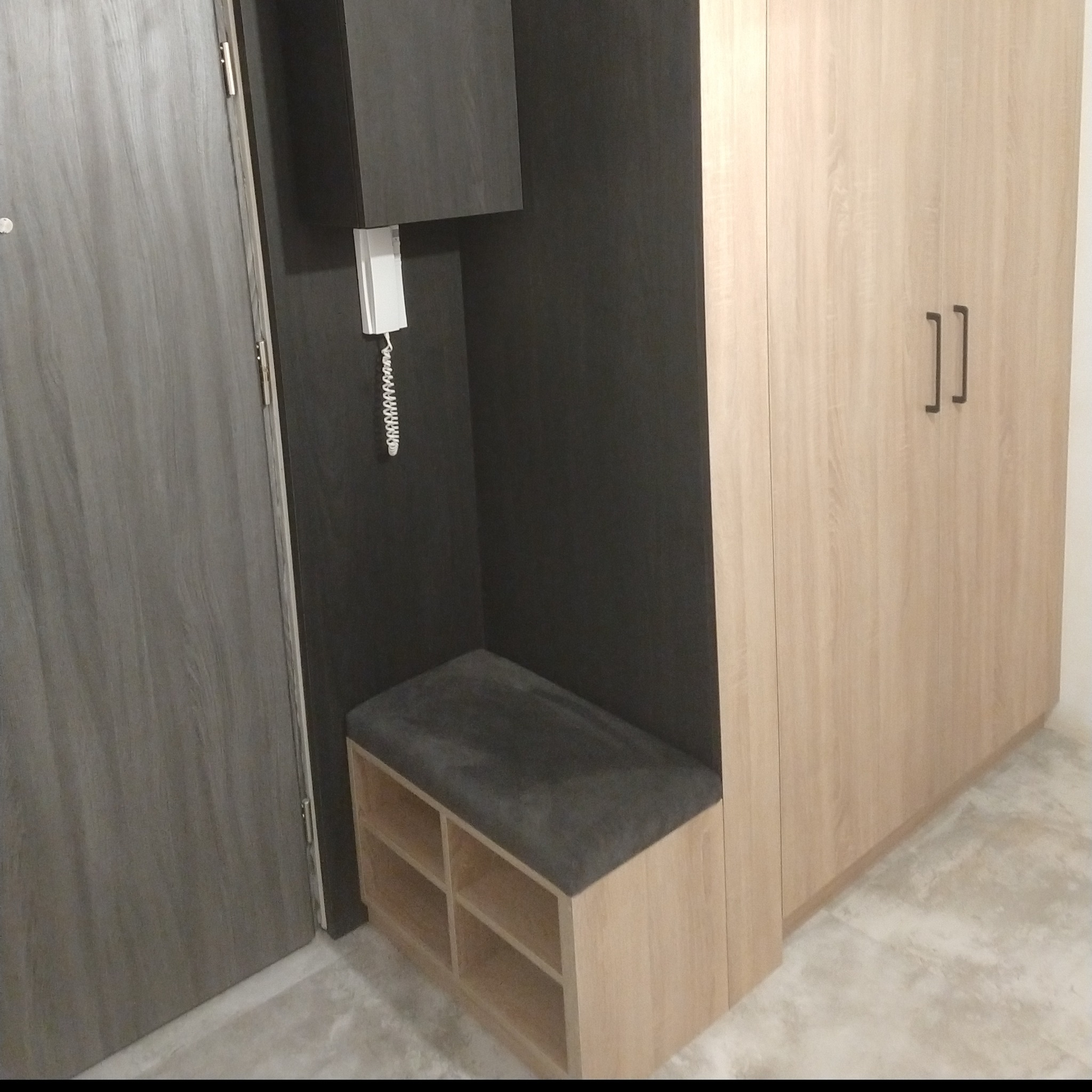 Wnęka z siedziskiem i półkami na buty, obok szafa z jasnego drewna, przy szarej ścianie z domofonem.