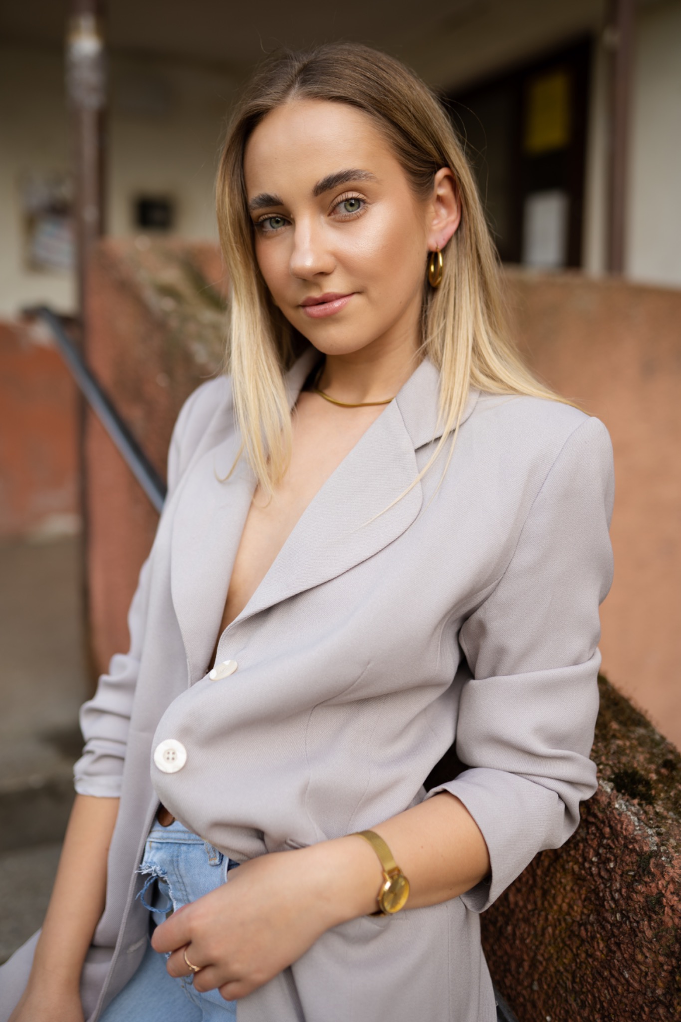 Portret młodej kobiety z blond włosami i zielonymi oczami, ubrana w szary blezer, złoty naszyjnik i zegarek, opierająca się o kamienne schody.