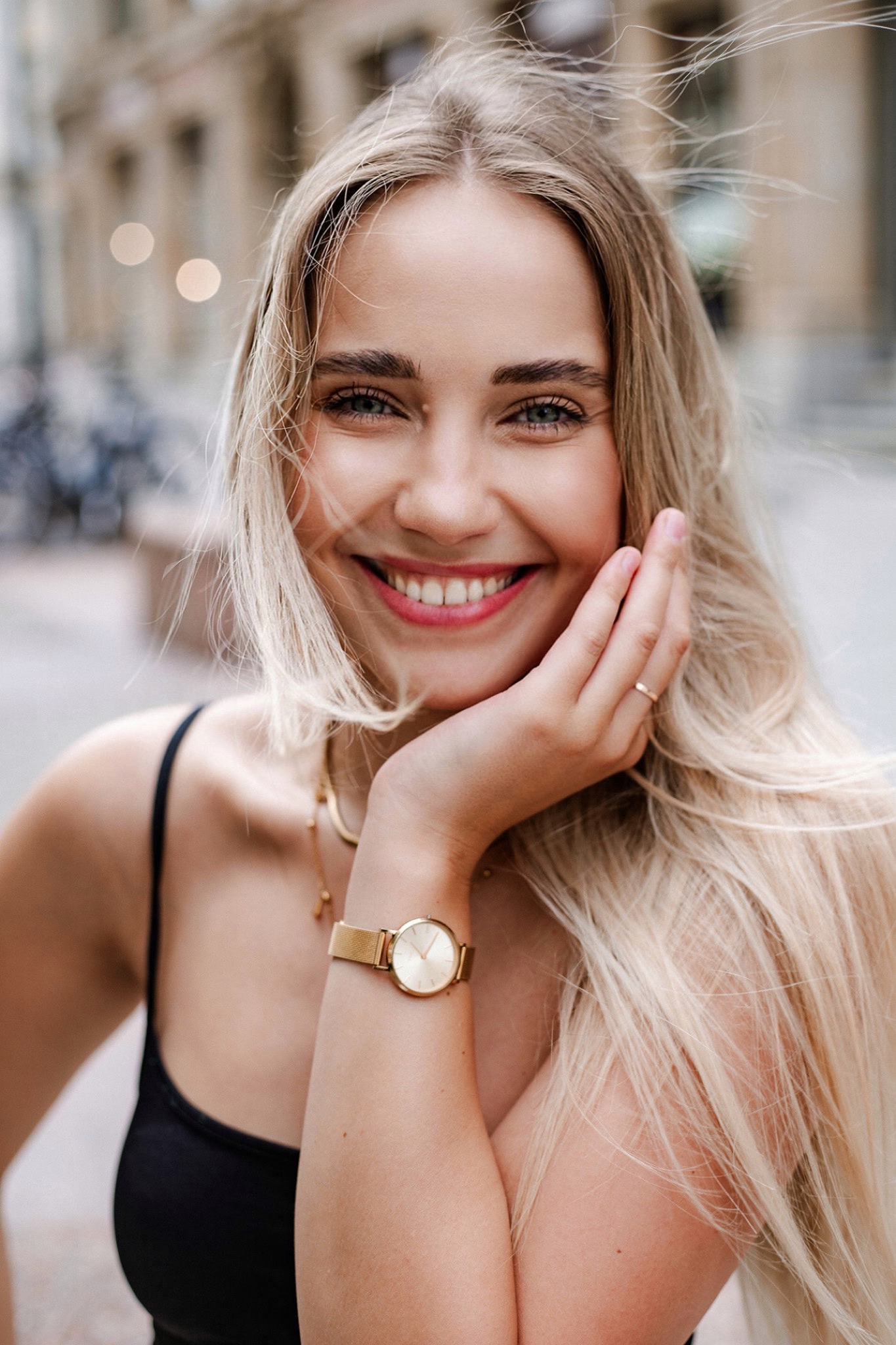 Uśmiechnięta blondynka w czarnym topie, z delikatnym makijażem i złotym zegarkiem na nadgarstku, trzymająca dłoń przy twarzy na tle miejskiej zabudowy.