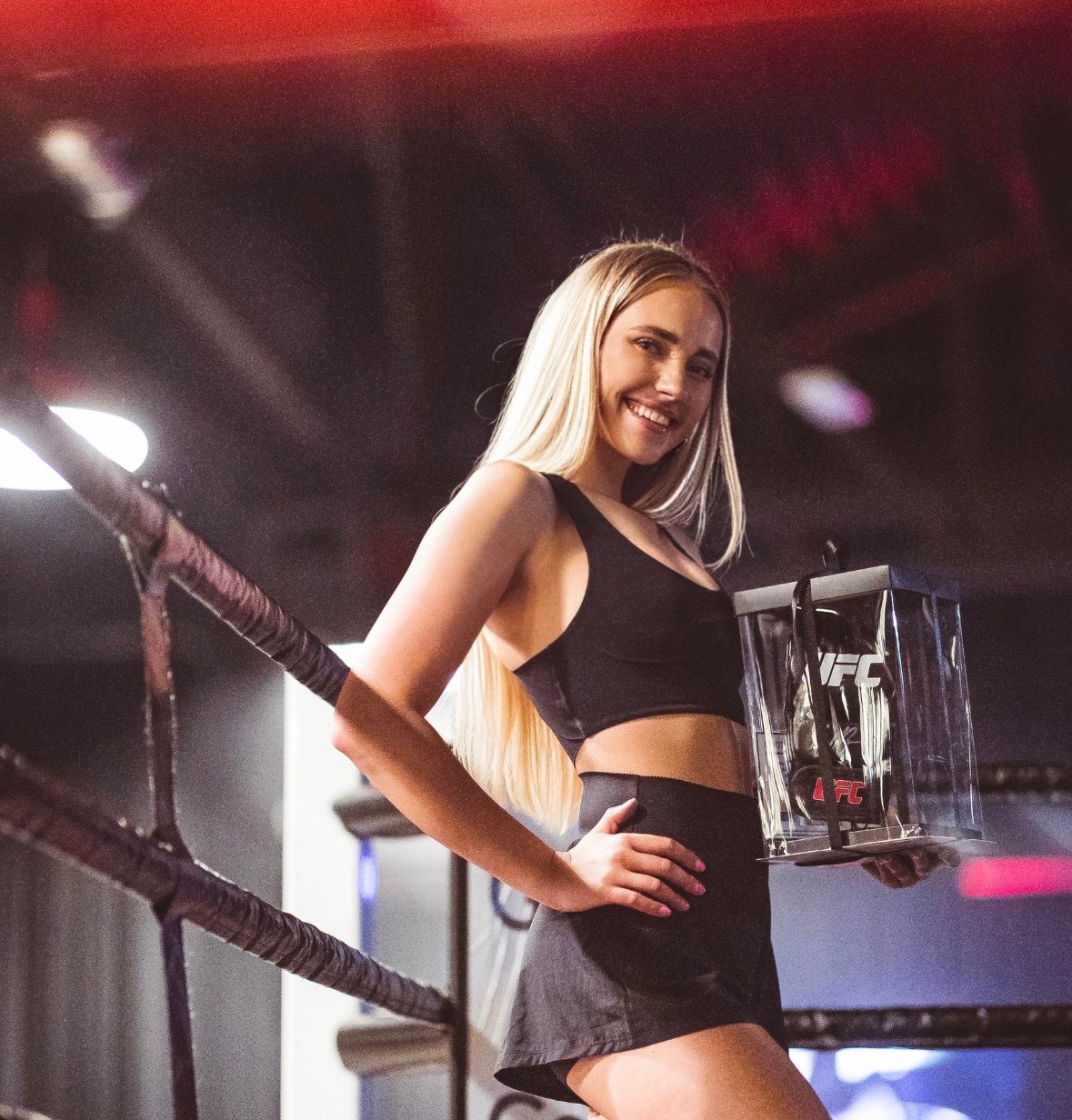 Uśmiechnięta blondynka w ringu bokserskim, trzymająca przezroczyste pudełko z logo UFC, spoglądająca w górę.