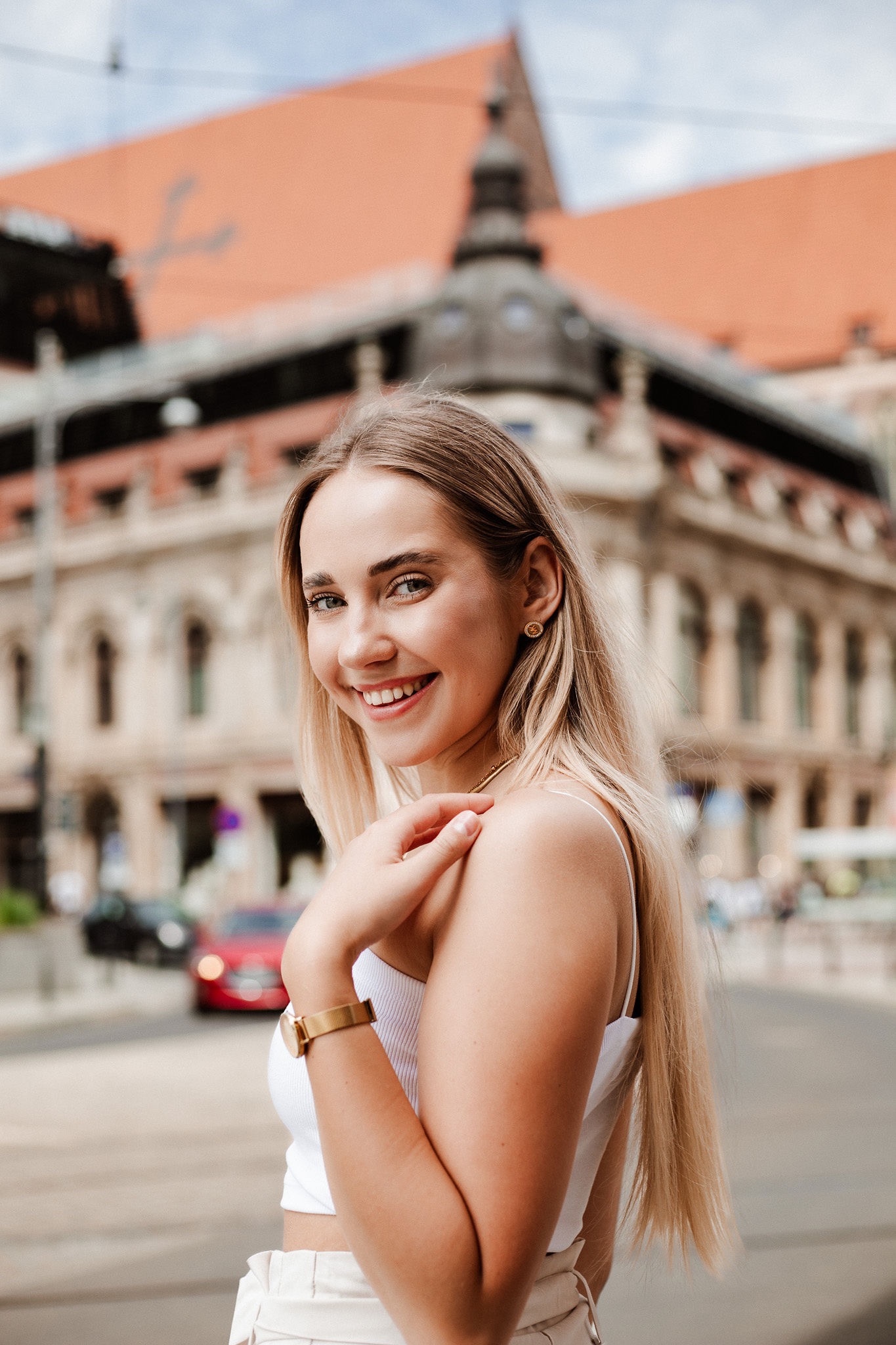 Portret uśmiechniętej blondynki w białym topie, złoty zegarek i kolczyki, na tle miejskiej architektury z czerwonym dachem