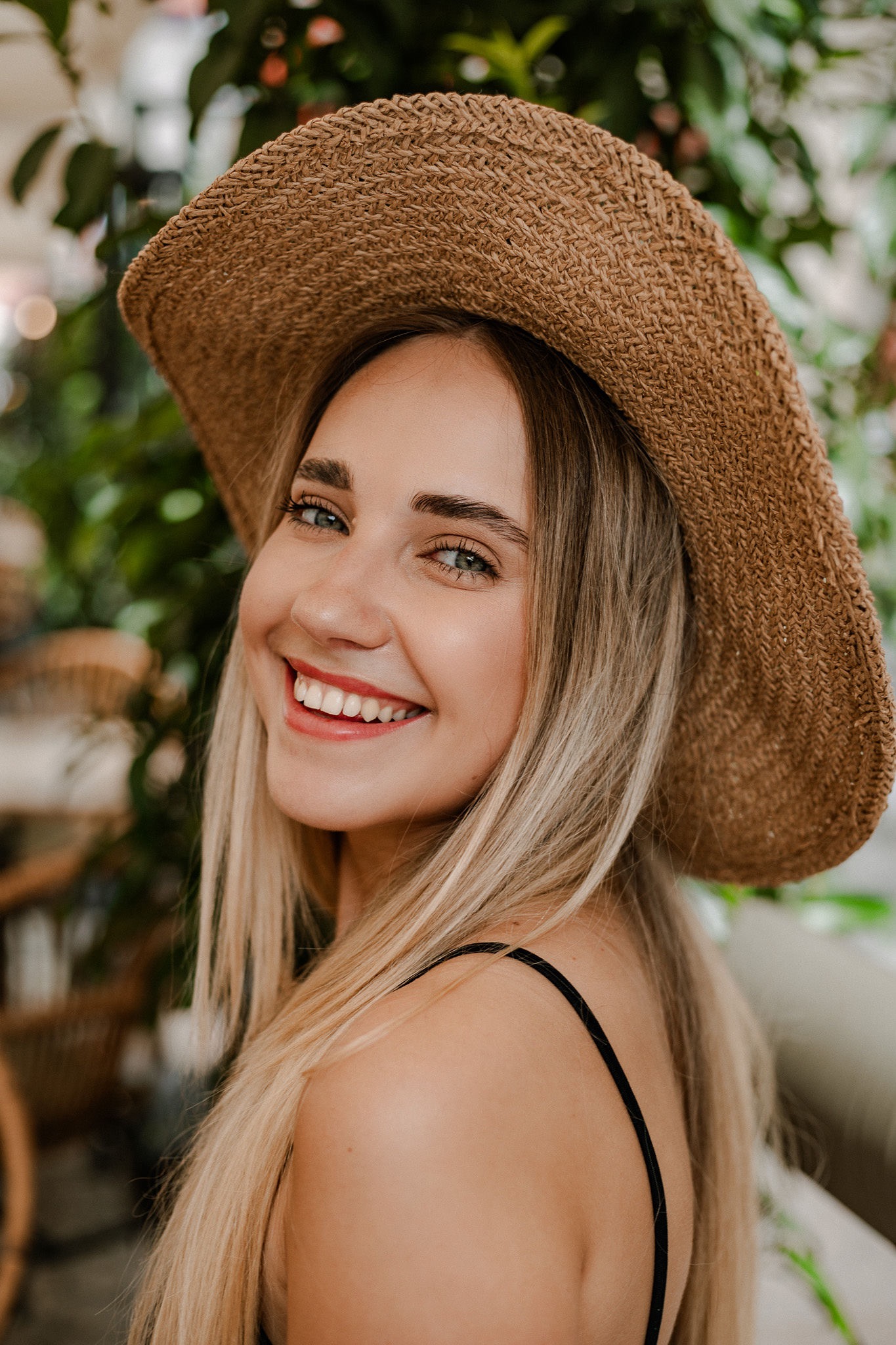 Uśmiechnięta blondynka w słomkowym kapeluszu, odwrócona przez ramię, na tle zieleni.