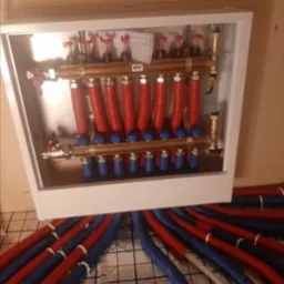 Rozdzielacz ogrzewania podłogowego w białej szafce z czerwono-niebieskimi rurami na folii aluminiowej z nadrukowaną siatką.