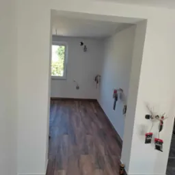 Wnętrze pokoju po remoncie, z panelami podłogowymi imitującymi drewno, świeżo pomalowanymi białymi ścianami i przygotowanymi punktami elektrycznymi, widoczne puszki po napojach energetycznych przy...