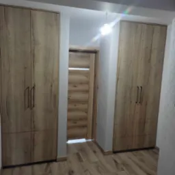 Dwie szafy na wymiar z drewnopodobnymi frontami, z czarnymi, długimi uchwytami, flankujące wejście z dopasowanymi kolorystycznie drzwiami z poziomym przeszkleniem, na podłodze panele w odcieniach...