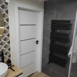 Nowoczesna łazienka z białymi drzwiami z czarnymi akcentami, grzejnikiem drabinkowym na szarej marmurowej ścianie i umywalką na drewnianym blacie.