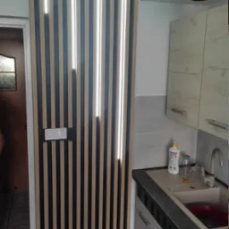 Drewniane lamele na ścianie z wbudowanym oświetleniem LED, obok fragmentu kuchni z umywalką i szafkami w jasnych odcieniach.