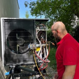Sławomir Kondej Kompleksowe systemy dla domu - Instalator w czerwonej koszulce serwisuje otwartą pompę ciepła na zewnątrz. Widoczne narzędzia i przewody, drzewa w tle. Pompa stoi na betonowej podstawie.