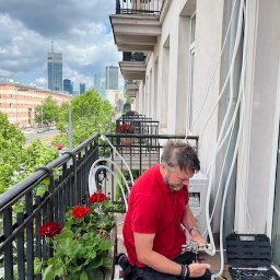 Sławomir Kondej Kompleksowe systemy dla domu - Montaż klimatyzacji na balkonie z widokiem na miasto. Instalator w czerwonej koszulce łączy przewody. Obok skrzynka z narzędziami i kwiaty w donicach.
