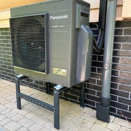 Sławomir Kondej Kompleksowe systemy dla domu - Szara pompa ciepła Panasonic Power Heat zamontowana na metalowym stelażu przy ścianie z czarnej cegły i drewnianym wykończeniem. Widoczna rura odprowadzająca.