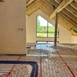 Instalacja ogrzewania podłogowego w nowym domu z drewnianą konstrukcją dachu. Rury grzewcze ułożone na folii termoizolacyjnej, widoczne okno w szczycie dachu.