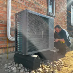 Instalacja pompy ciepła Panasonic przez montera na tle ceglanej ściany budynku z widocznymi przewodami i oknem. Podstawa pompy otoczona kamieniami.