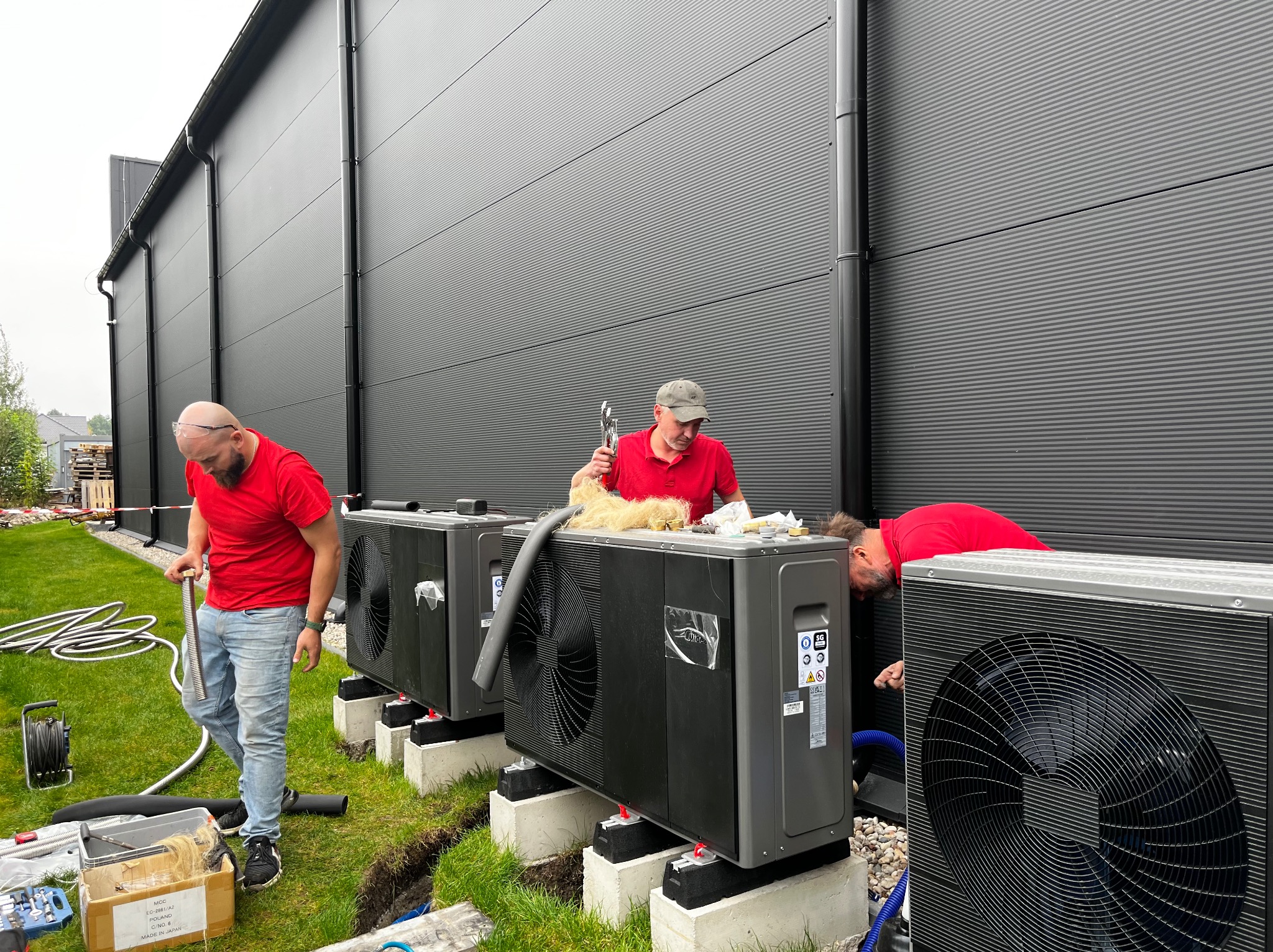Montaż trzech szarych pomp ciepła na betonowych podstawach, pracownicy w czerwonych koszulkach podczas instalacji na zewnątrz budynku z szarą elewacją.