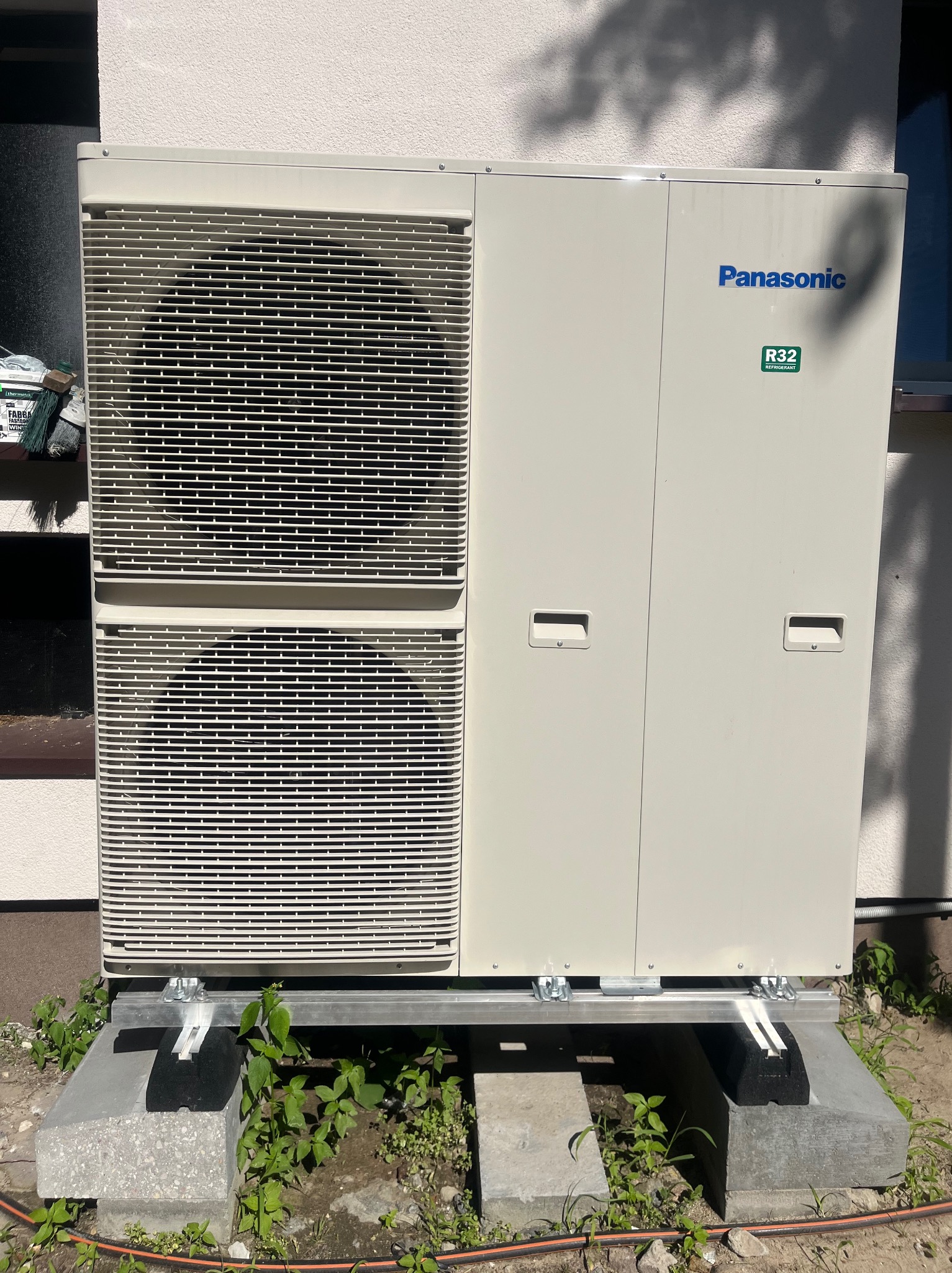 Zewnętrzna jednostka pompy ciepła Panasonic R32 na betonowej podstawie, z widocznymi elementami montażowymi. Obok ściana budynku i roślinność.
