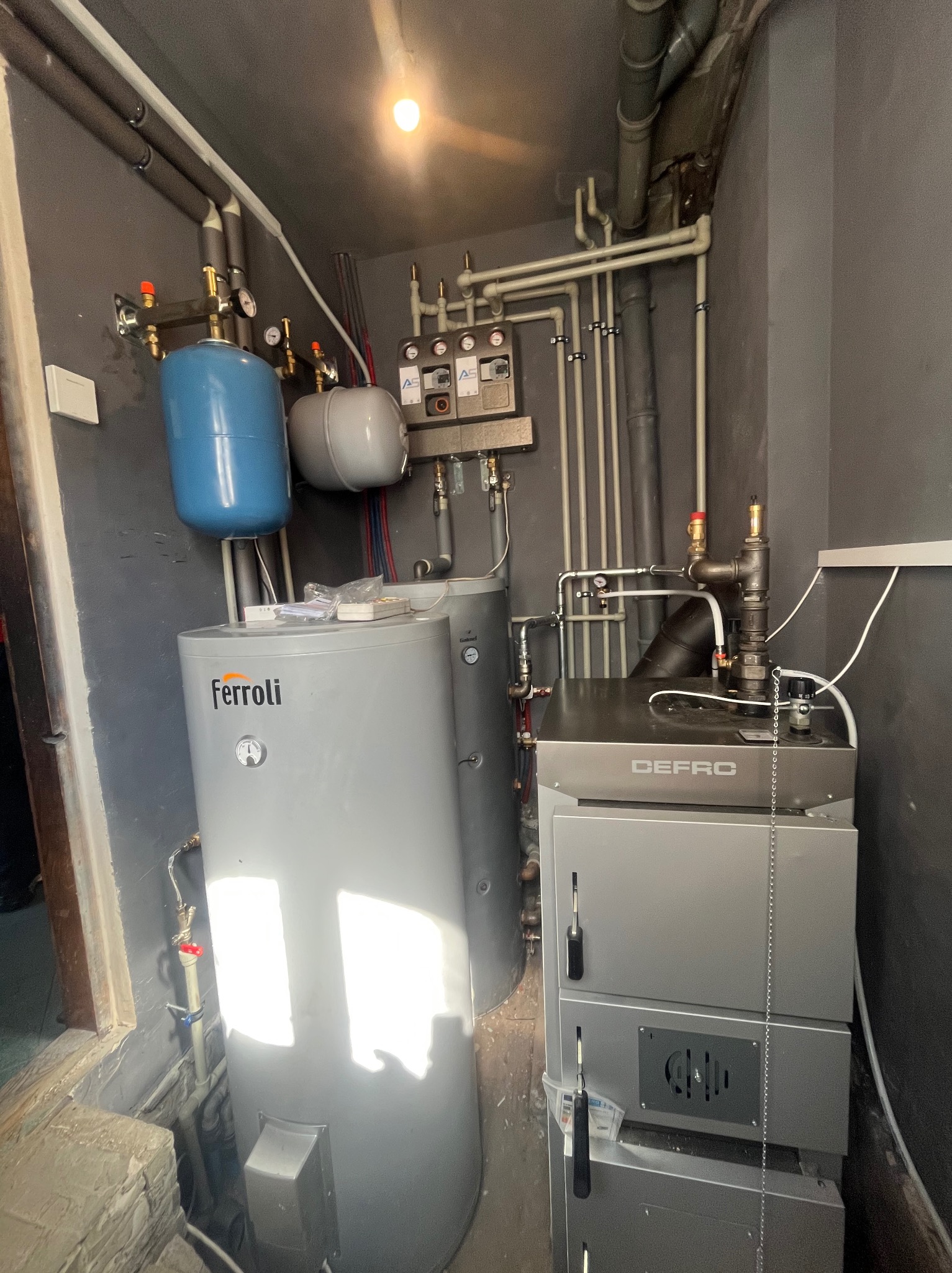 Kotłownia z piecem DEFRO i zasobnikiem Ferroli. Instalacja grzewcza w szarym pomieszczeniu z widocznymi rurami i elementami sterującymi.