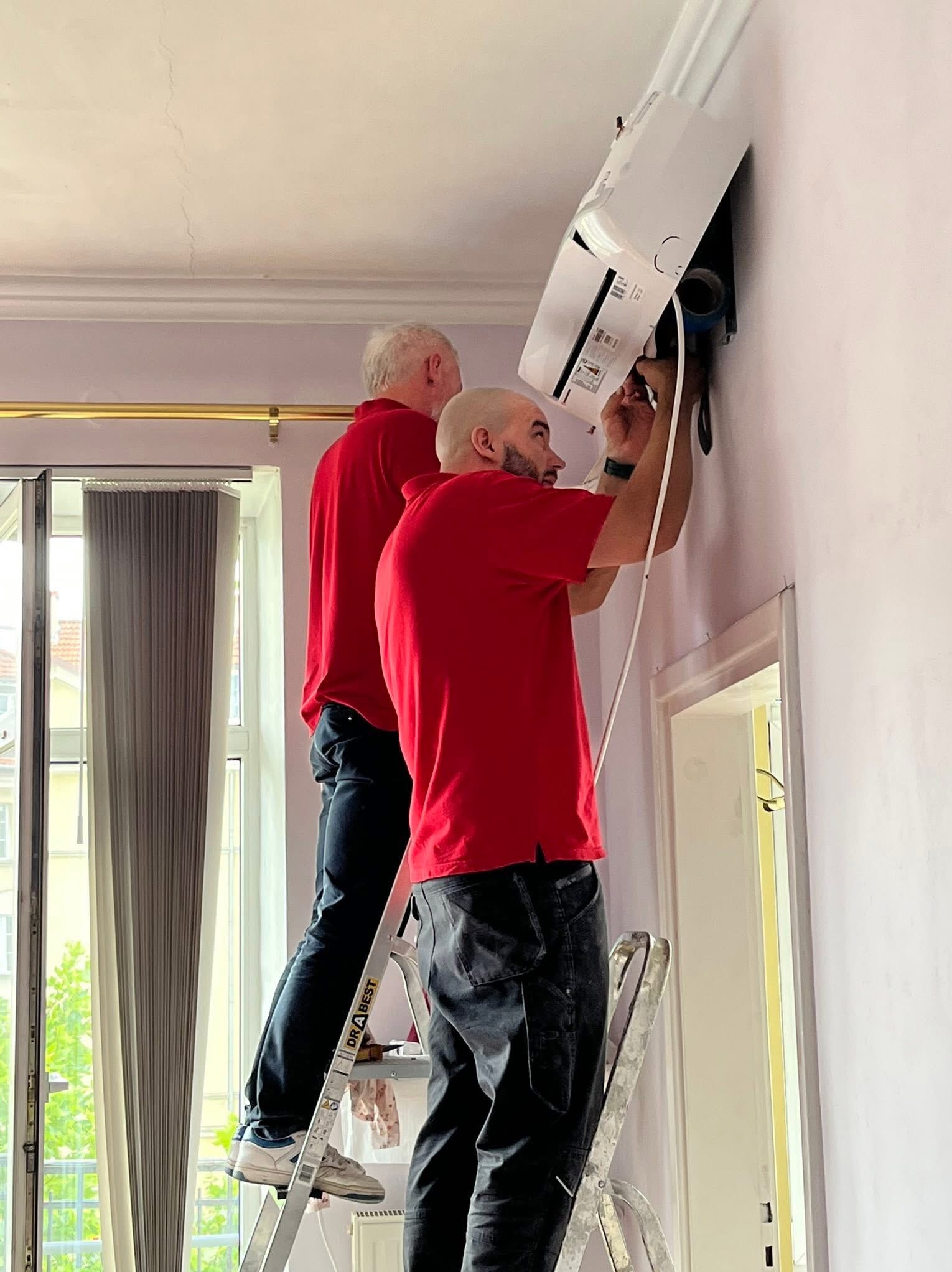 Montaż klimatyzacji w domu w Nowym Żabieńcu. Dwóch monterów na drabinie podłącza biały klimatyzator ścienny w pomieszczeniu z oknem i ozdobną listwą sufitową.