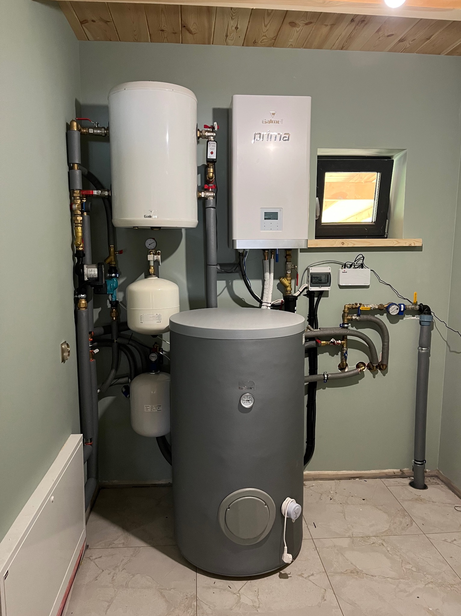 Kompletna instalacja grzewcza z szarym zbiornikiem akumulacyjnym, białym zasobnikiem CWU i kotłem gazowym w pomieszczeniu z drewnianą boazerią.