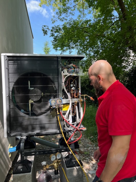 Instalator w czerwonej koszulce serwisuje otwartą pompę ciepła na zewnątrz. Widoczne narzędzia i przewody, drzewa w tle. Pompa stoi na betonowej podstawie.
