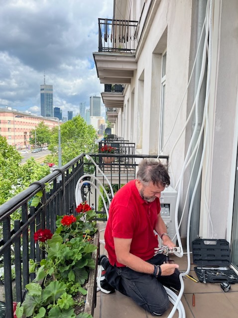 Montaż klimatyzacji na balkonie z widokiem na miasto. Instalator w czerwonej koszulce łączy przewody. Obok skrzynka z narzędziami i kwiaty w donicach.