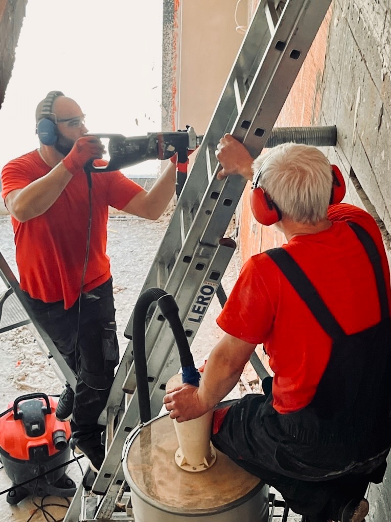 Dwóch monterów w czerwonych koszulkach wierci otwór w ścianie za pomocą wiertnicy z odsysaniem pyłu, stojąc na drabinie. Praca w słuchawkach ochronnych.