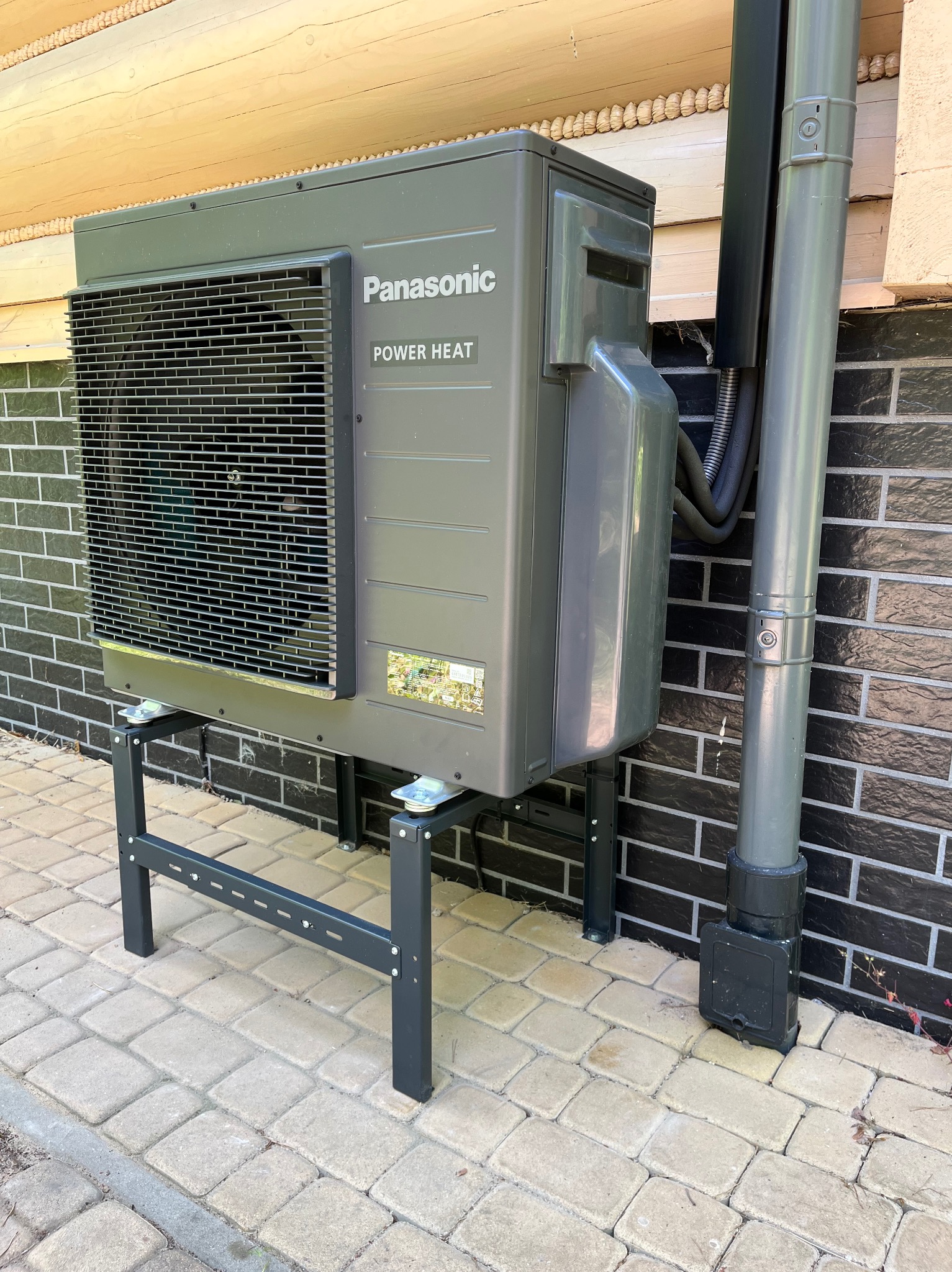 Szara pompa ciepła Panasonic Power Heat zamontowana na metalowym stelażu przy ścianie z czarnej cegły i drewnianym wykończeniem. Widoczna rura odprowadzająca.
