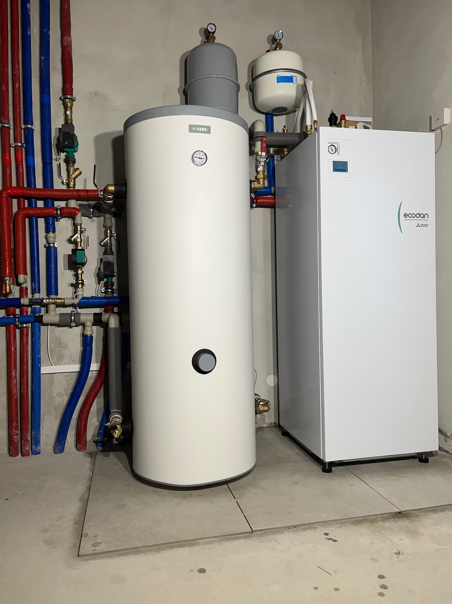 Biały zasobnik ciepłej wody NIBE i pompa ciepła Ecodan Mitsubishi z kompletną instalacją hydrauliczną w pomieszczeniu technicznym.