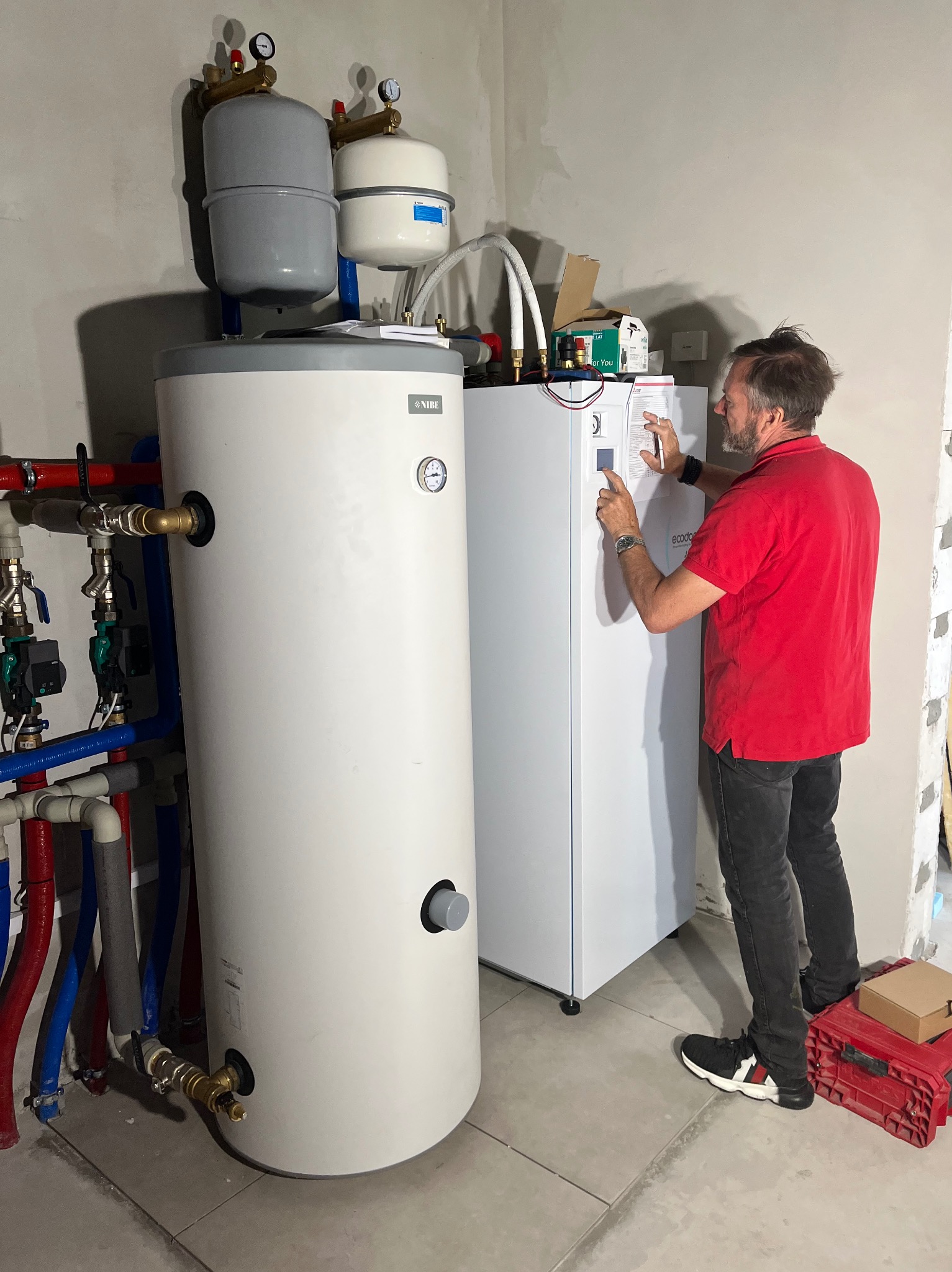 Instalator w czerwonej koszulce konfiguruje pompę ciepła NIBE w surowym pomieszczeniu, widoczne rury i zbiorniki. Na podłodze płytki i skrzynka z narzędziami.