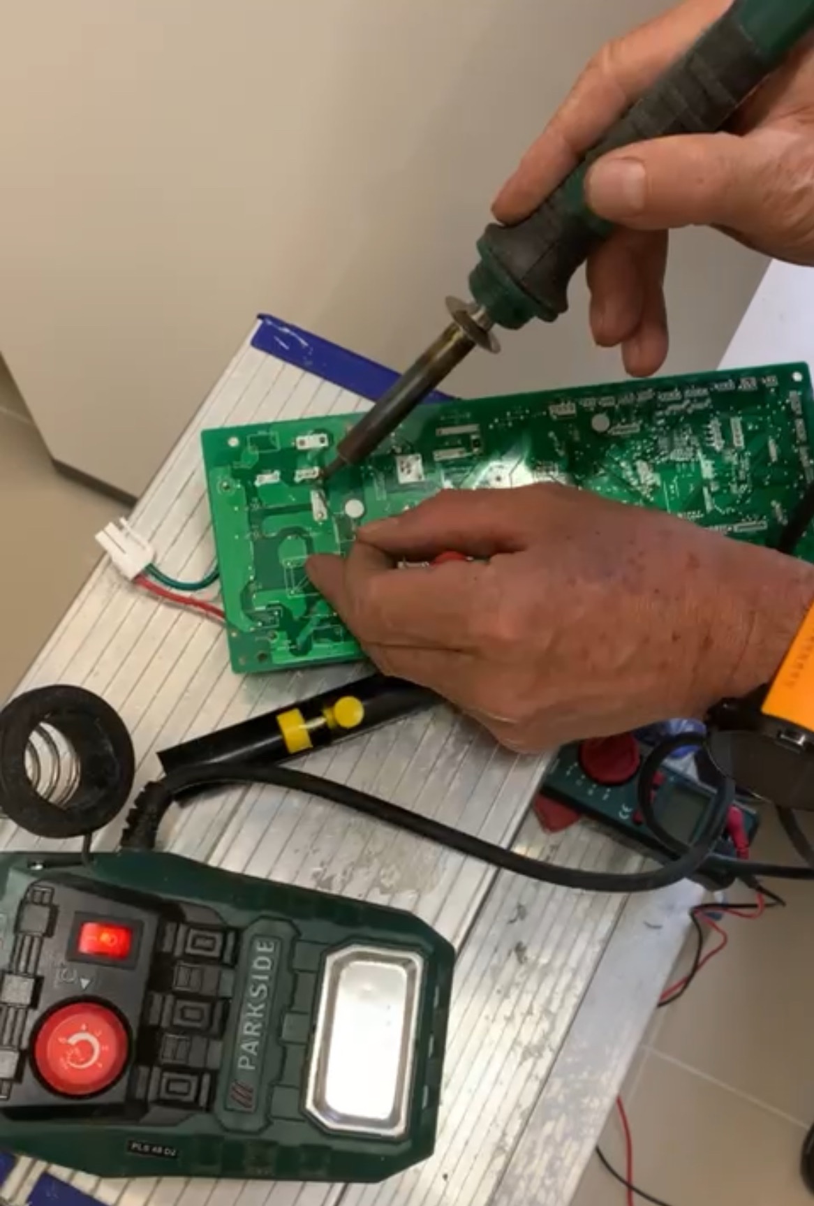 Naprawa elektroniki: lutowanie zielonej płytki drukowanej przez serwisanta, widoczne narzędzia Parkside, miernik, kabel i lutownica na metalowej drabinie.