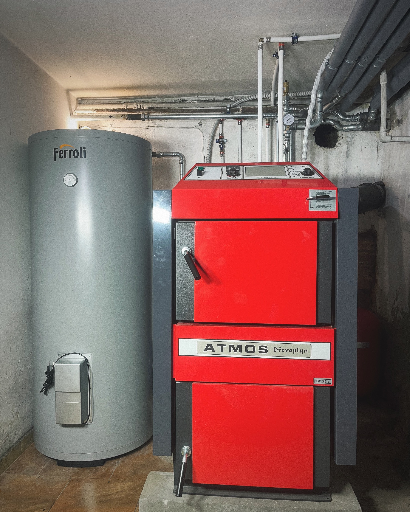 Czerwono-szary piec Atmos Drevo plyn obok szarego zbiornika Ferroli w kotłowni. Widoczne rury i instalacje na ścianie. Oświetlenie górne.