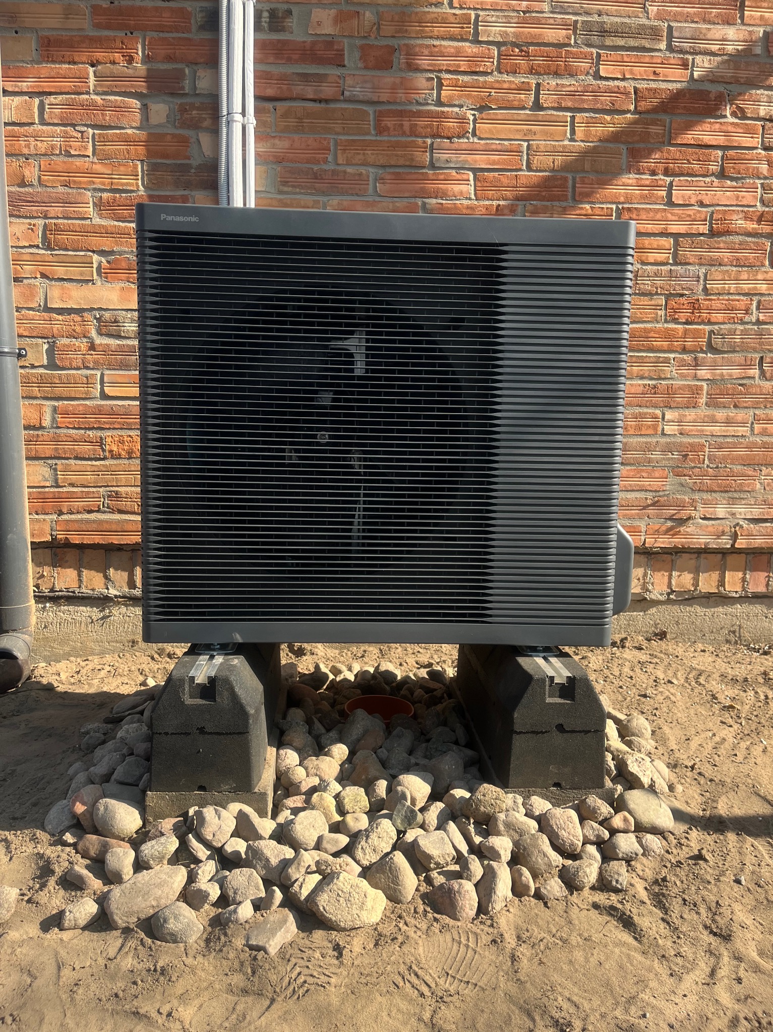 Szara pompa ciepła Panasonic, zainstalowana na betonowych blokach i otoczona kamieniami, na tle ceglanej ściany. Widoczne rury pionowe.