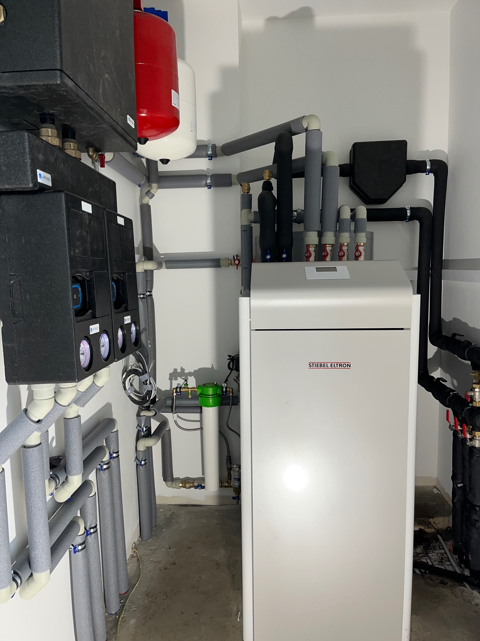 Biała pompa ciepła Stiebel Eltron z podłączonymi rurami, zaworami i zbiornikami w pomieszczeniu technicznym. Szare izolacje na rurach.
