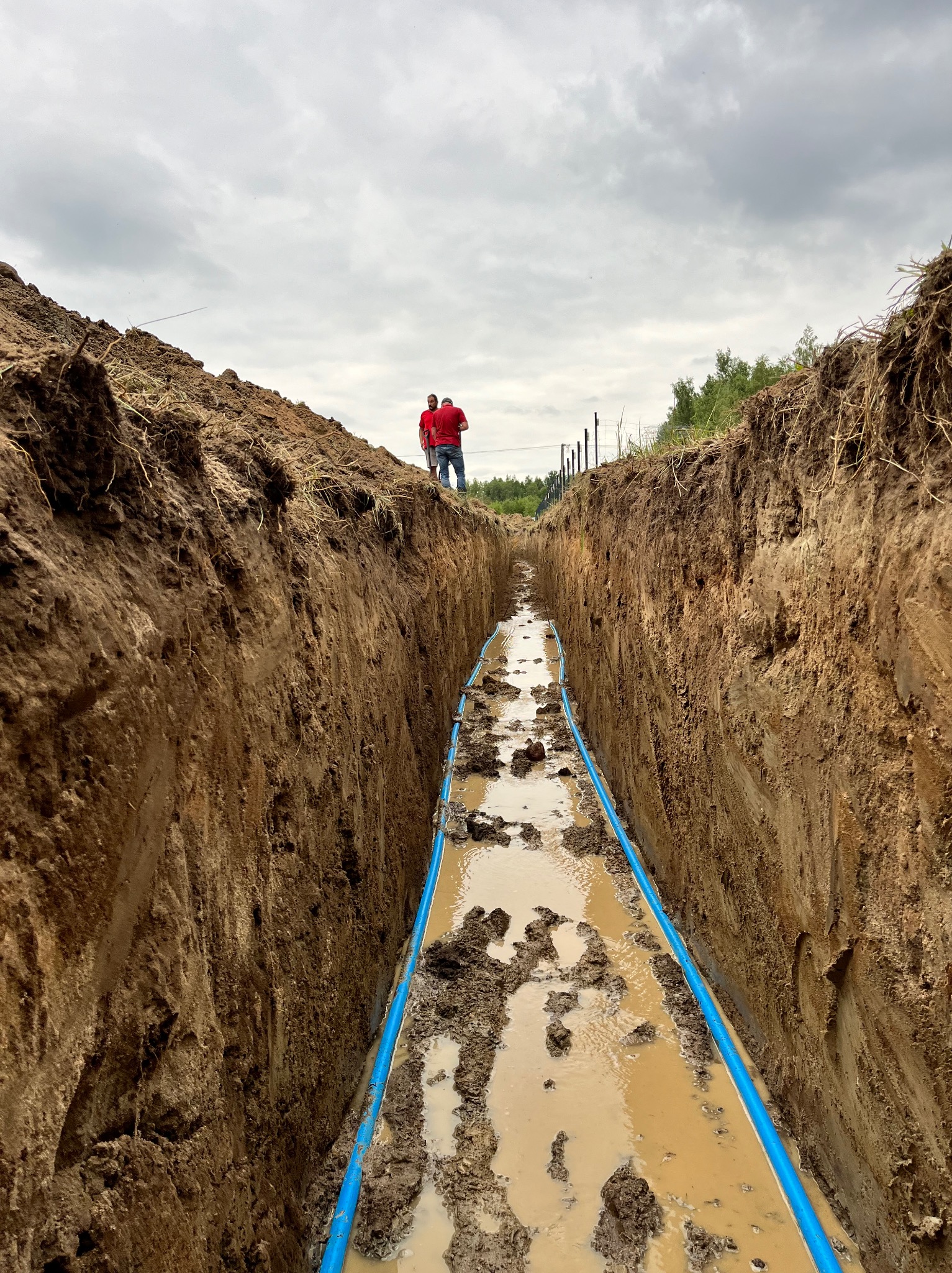 Wykop z ułożonym niebieskim rurociągiem w błotnistym terenie. Dwóch mężczyzn w czerwonych koszulkach obserwuje z góry. Pochmurne niebo w tle.