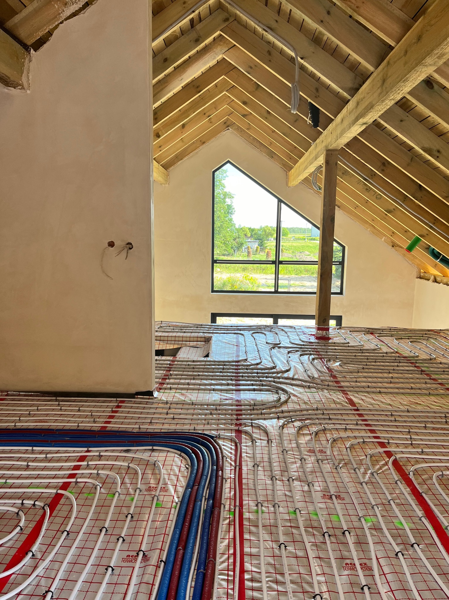 Instalacja ogrzewania podłogowego w nowym domu z drewnianą konstrukcją dachu. Rury grzewcze ułożone na folii termoizolacyjnej, widoczne okno w szczycie dachu.