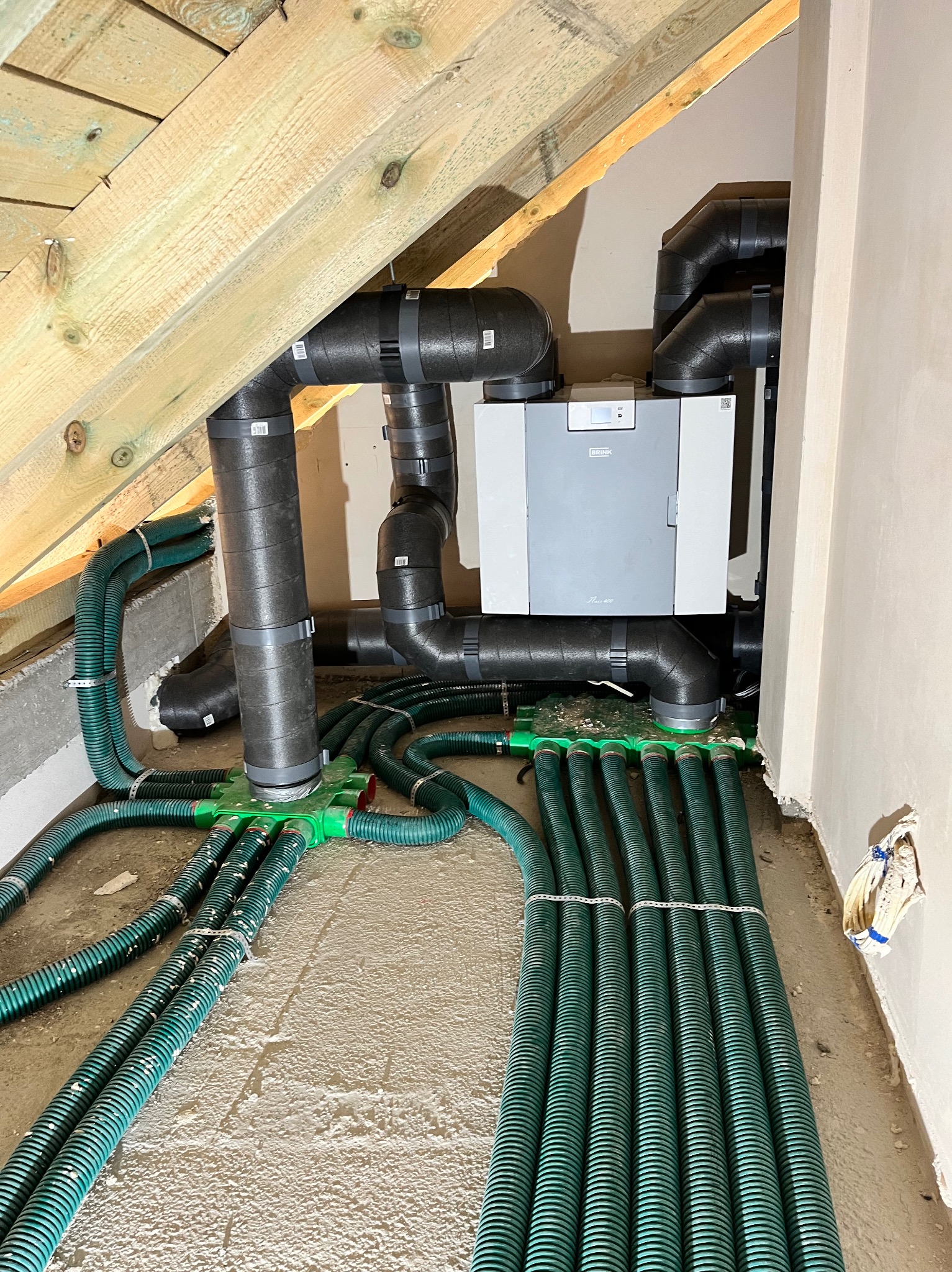 System rekuperacji Brink w przestrzeni poddasza: szare urządzenie, czarne rury izolowane i zielone przewody wentylacyjne na surowym podłożu. Widoczne elementy konstrukcji dachu.