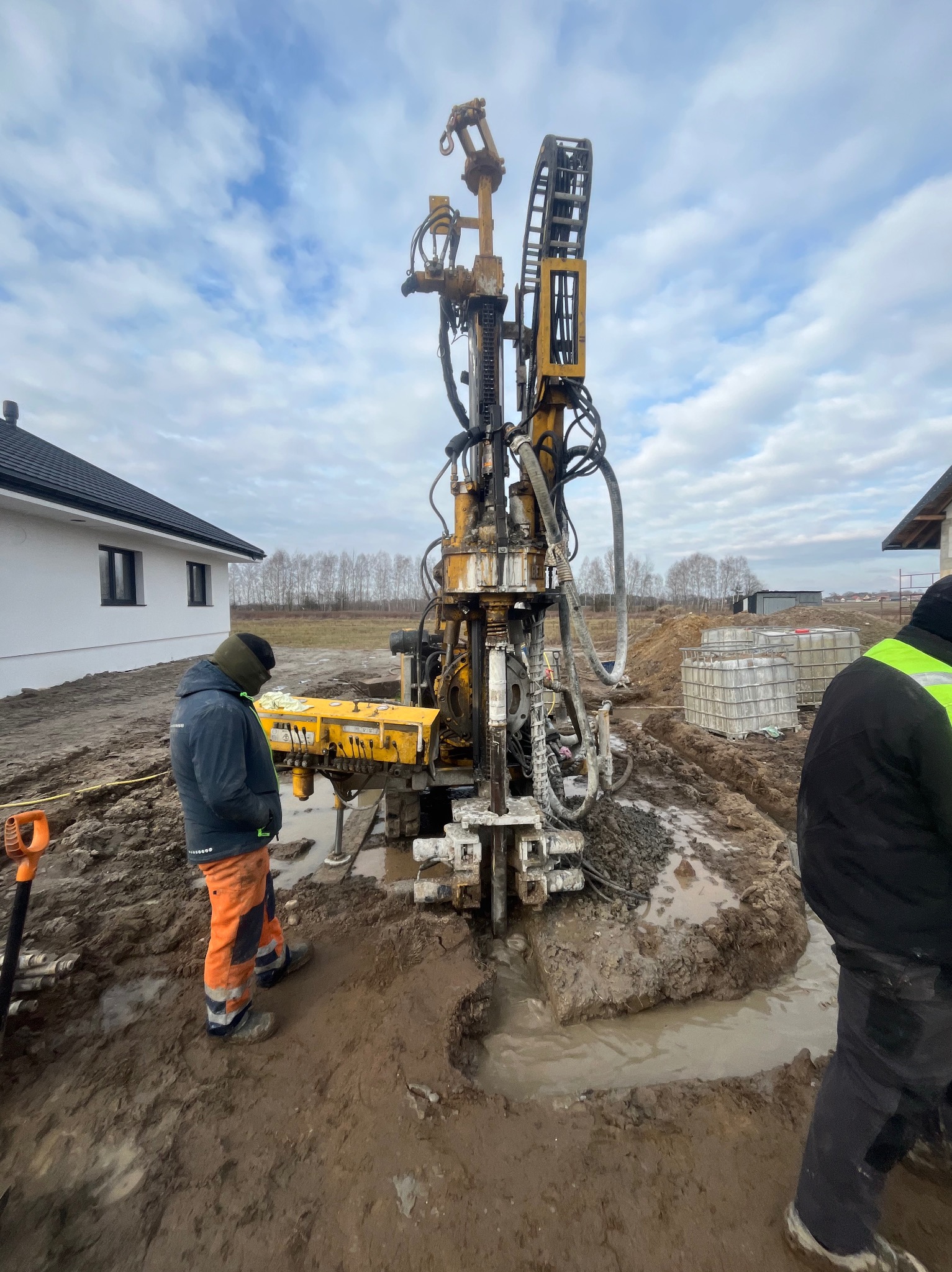 Wiertnica w trakcie pracy przy budowie domu. Widoczne błoto, pracownicy i fragment budynku. Pochmurne niebo w tle.