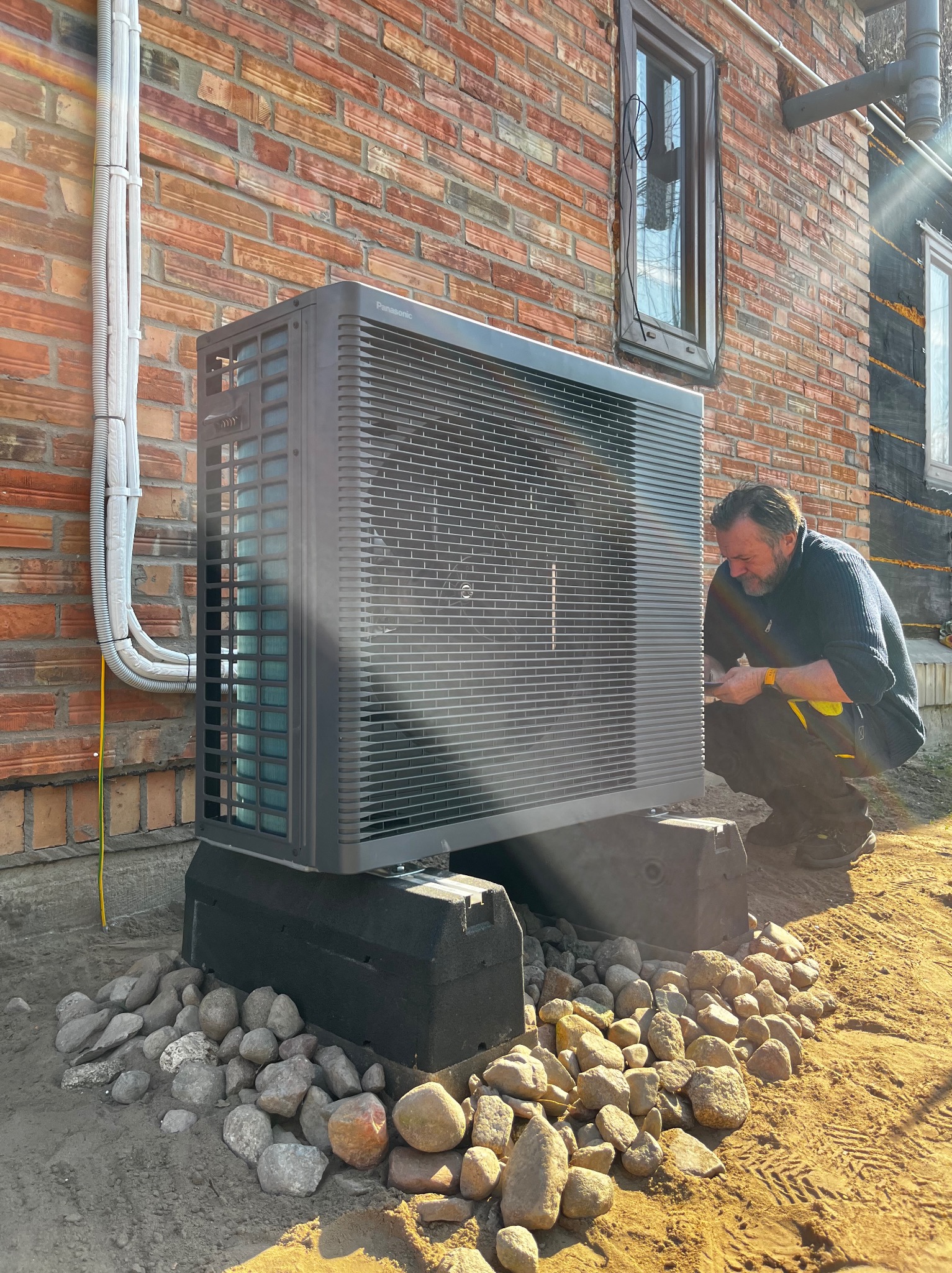 Instalacja pompy ciepła Panasonic przez montera na tle ceglanej ściany budynku z widocznymi przewodami i oknem. Podstawa pompy otoczona kamieniami.