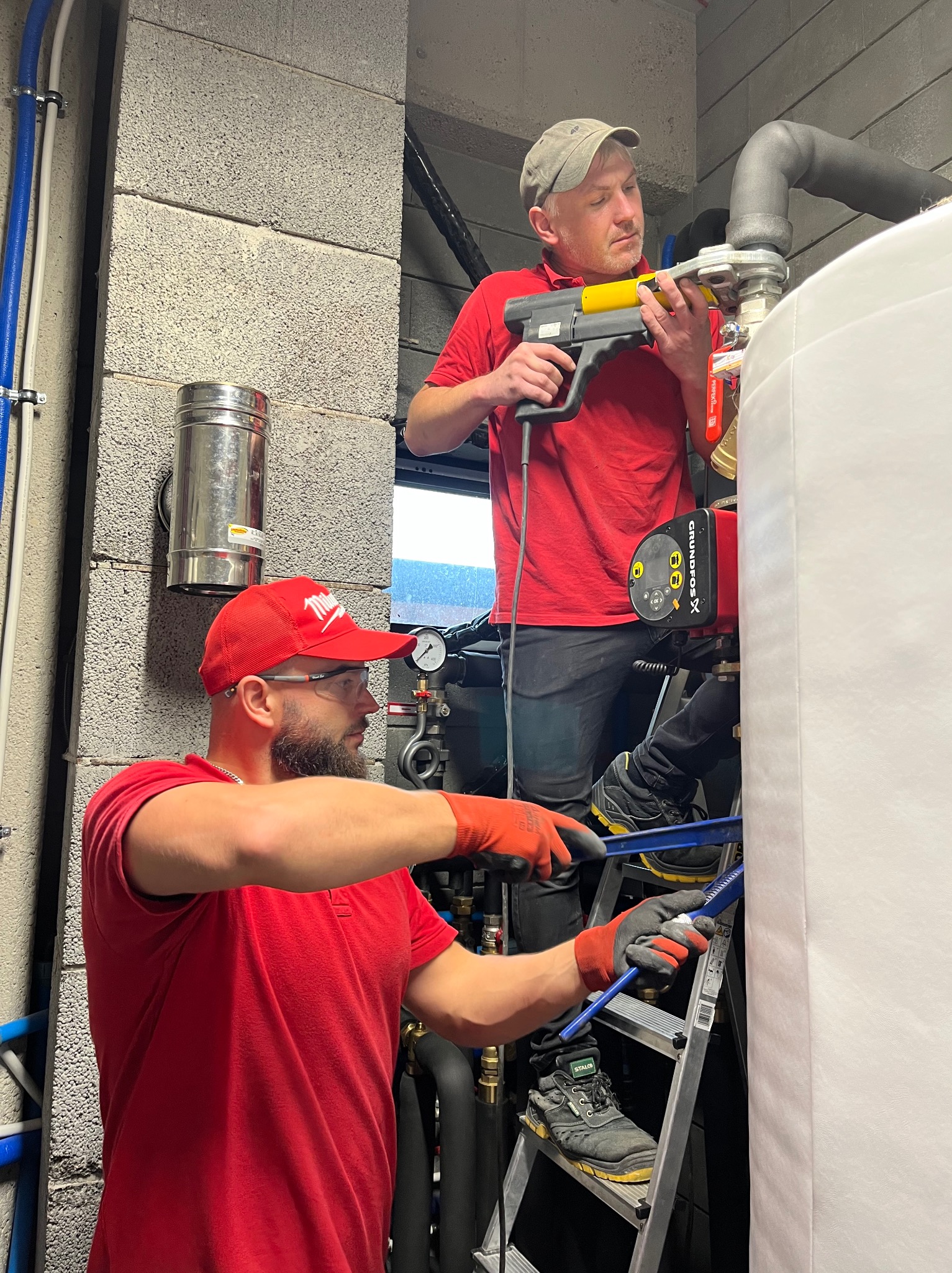 Dwóch hydraulików w czerwonych koszulkach łączy rury przy użyciu specjalistycznych narzędzi na tle szarej ściany z bloków i białego zbiornika. Jeden stoi na drabinie.