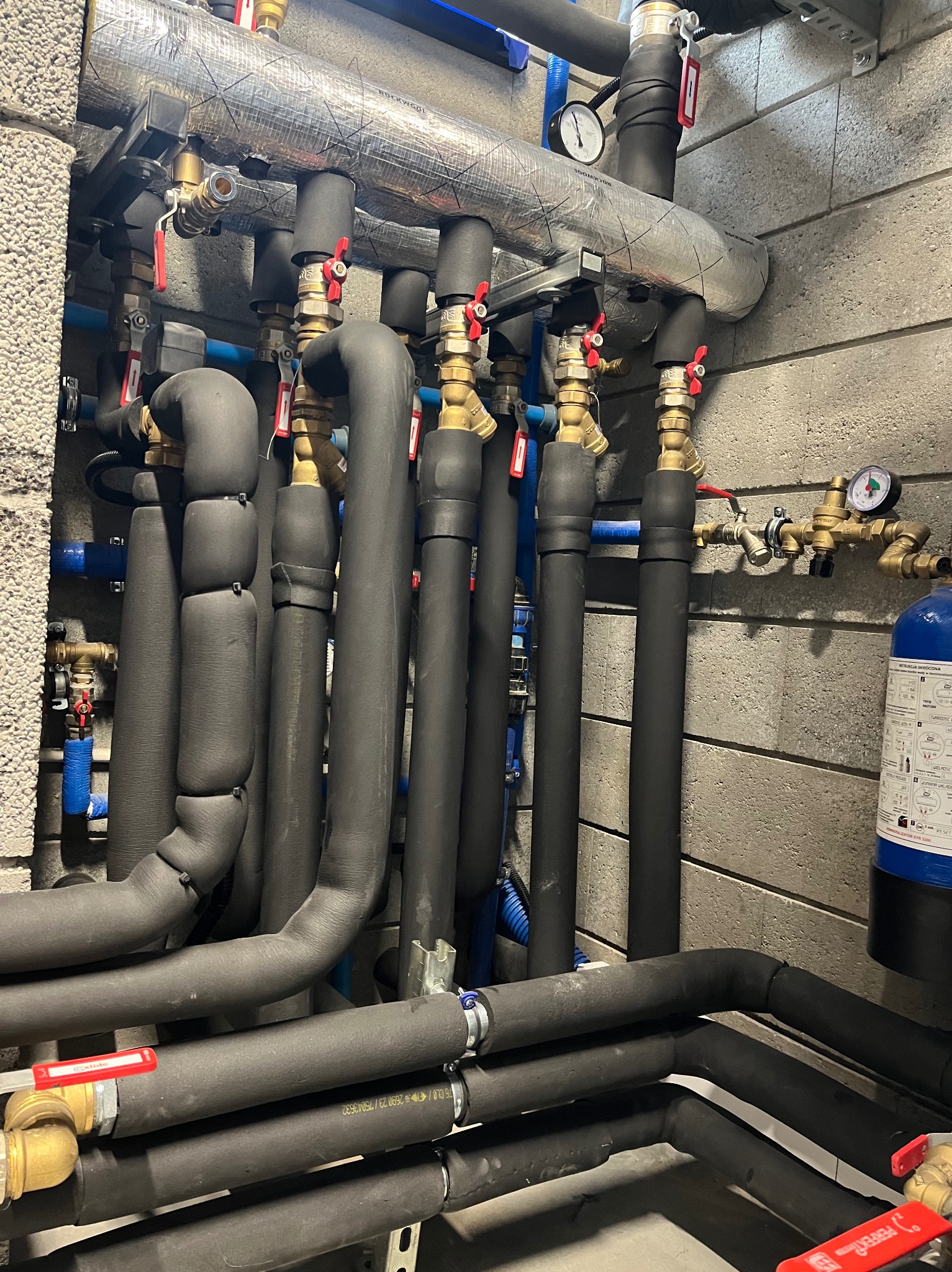 Złożona instalacja hydrauliczna z rurami w izolacji, zaworami z czerwonymi uchwytami i manometrami na tle betonowej ściany. Widoczne elementy mocujące i niebieskie rury.