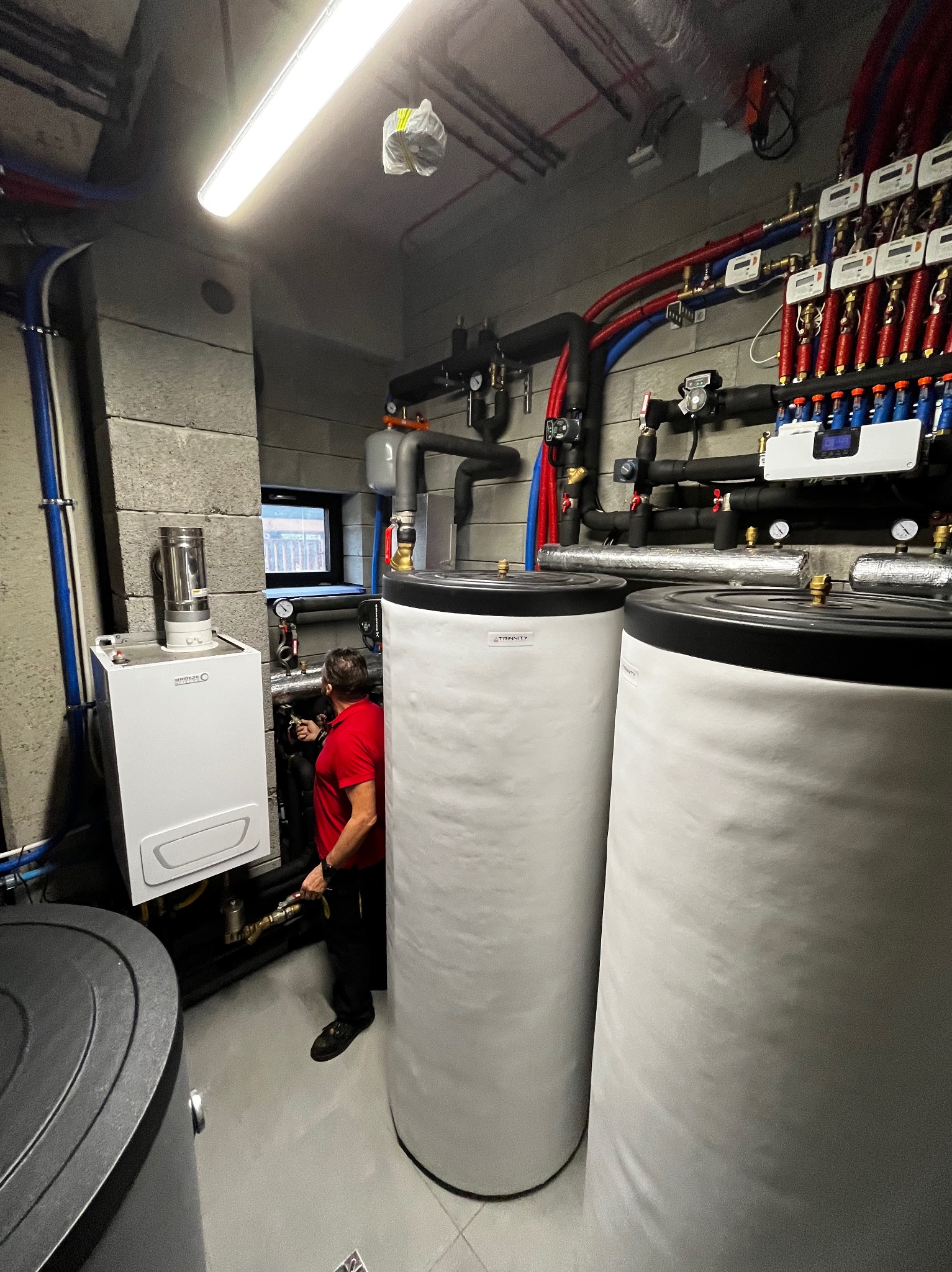 W kotłowni: biały piec gazowy, dwa białe zbiorniki akumulacyjne, instalacja z rur i zaworów. Mężczyzna w czerwonej koszulce przy piecu. Szare ściany z betonu.