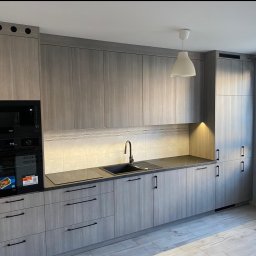 Włodarczyk Meble - Sandra Włodarczyk - Jasnoszara kuchnia na wymiar z czarnymi uchwytami i sprzętem AGD. Uwagę zwraca minimalistyczny design i oświetlenie podszafkowe.